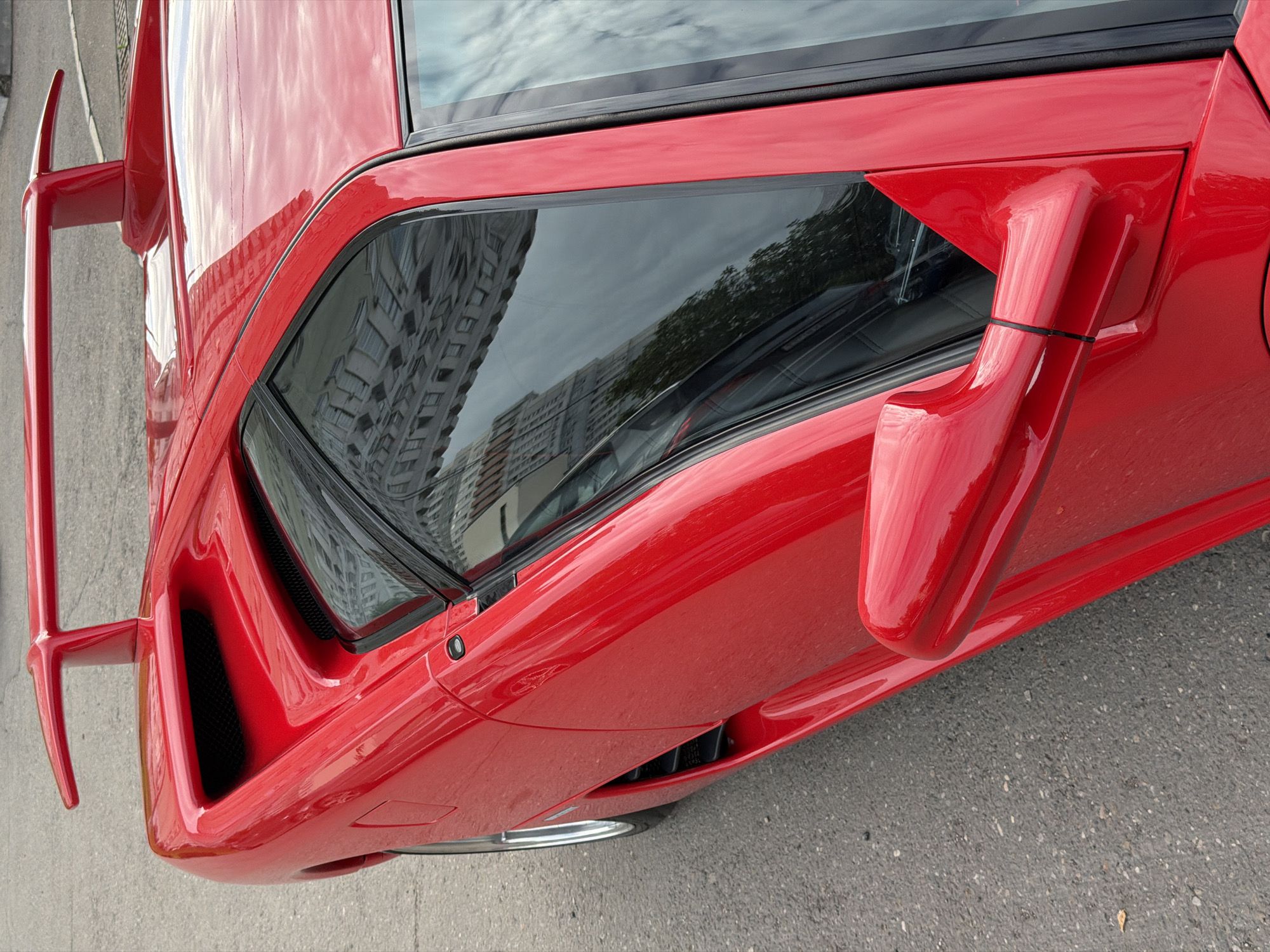 An elevated, close-up view captures a vibrant red supercar, identifiable as a Lamborghini Countach, parked outdoors on an asphalt surface. The car's glossy red paint reflects the surrounding environment.
The angular bodywork of the classic supercar is prominent, showcasing its distinctive design features. On the passenger side, a large, triangular side mirror is mounted, secured at its base with a black zip tie. Further back, a deeply recessed side air intake with a black mesh grille is visible. On the driver's side, a unique, periscope-like mirror stalk, characteristic of early Countach models, extends upwards from the bodywork.
Reflections on the car's windows and highly polished paintwork reveal tall, multi-story buildings, likely residential or commercial apartment blocks, as well as a cloudy sky and hints of trees. These reflections indicate an urban setting. The lighting suggests it is daytime, possibly overcast.
The vehicle appears to be stationary, with no human activity depicted. The asphalt ground is visible beneath the car. There is no visible text in the image. The location context for this scene is Moscow, Russia.