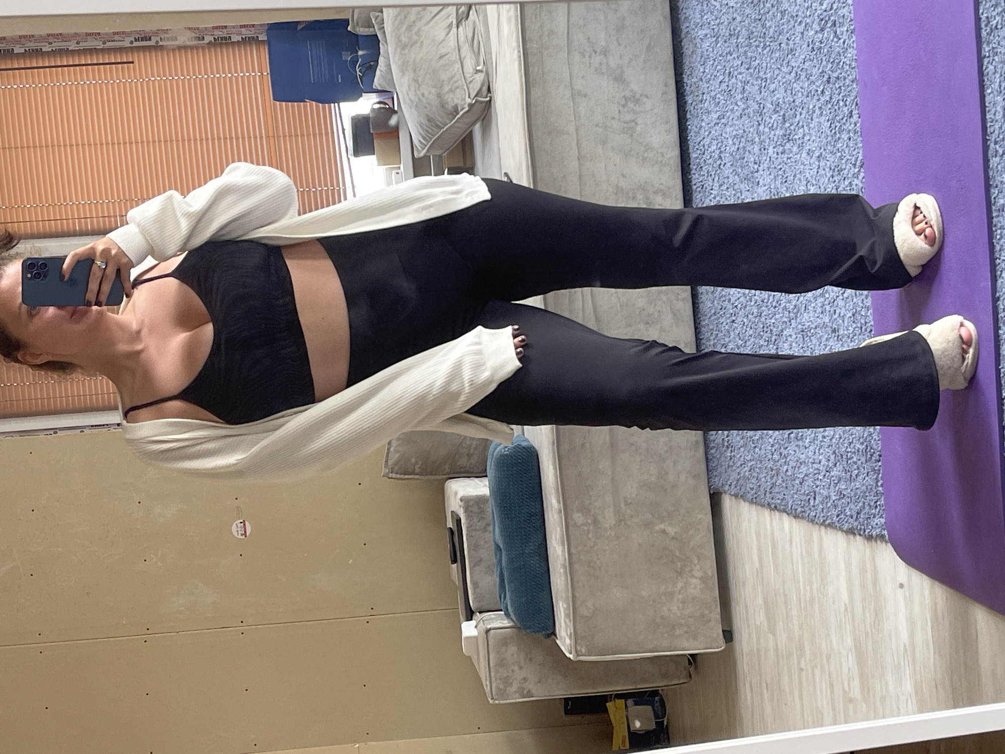 A woman is seen in a mirror selfie, wearing a black sports bra, black pants, and a white cardigan. She holds a blue phone. She's wearing fuzzy slippers. The background includes a window with blinds, a couch with pillows, a purple yoga mat, and a beige wall. The location is unclear and not ascertainable from the image.