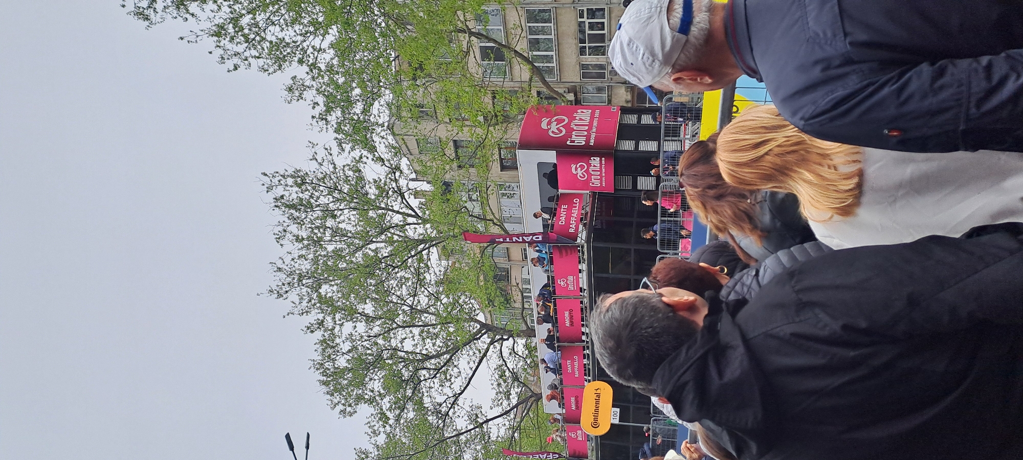 The image shows a crowd of people gathered near a promotional structure or stage in Burgas, Bulgaria. The stage is branded with "Giro d'Italia" logos, suggesting an event related to the cycling race. Banners with "Dante Raffaello" and "Amore Infinito" are also visible. The crowd is diverse in age and attire. The backdrop includes a building and trees, typical of an urban environment. A prominent detail is a "Continental" sign, likely representing a sponsor of the event.