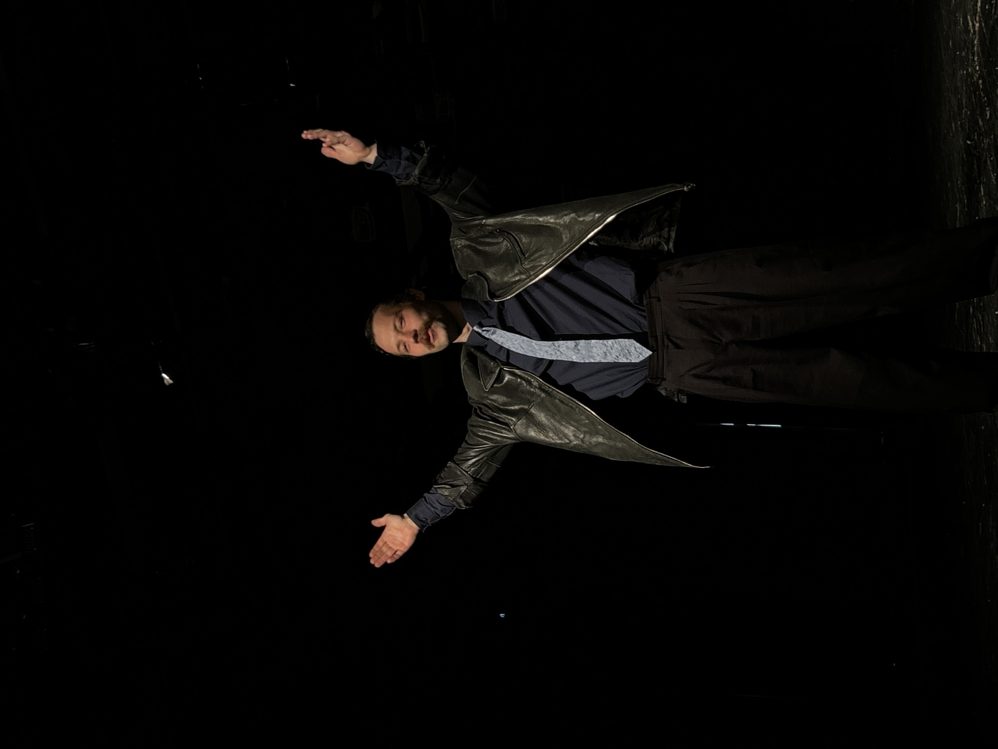 A man is standing on a stage. He is wearing a navy shirt, a light blue tie, black pants, and a black leather jacket. He has a beard and is smiling. He is standing with his arms outstretched. The background is black. The location is unknown.