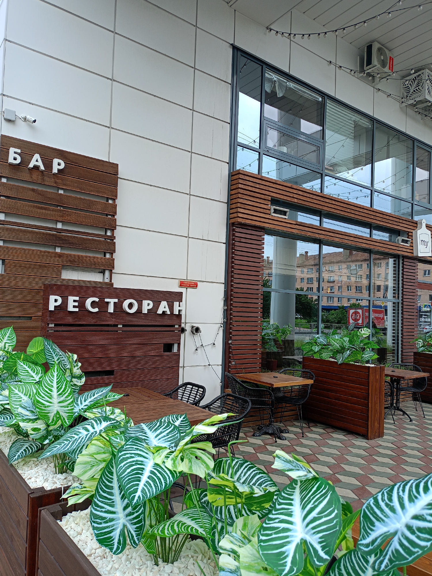 The image shows the exterior of a restaurant and bar. The building has a modern design with a white tiled facade and large windows framed by dark wood. Outside the establishment, there are several tables and chairs set up on a paved patio. Green plants in wooden planters add a touch of nature to the scene.
The words "БАР" (BAR) and "РЕСТОРАН" (RESTAURANT) are prominently displayed on wooden signs, indicating the establishment's dual function. Above the restaurant entrance, a small sign with the word "my" and a white cutting board icon is visible.
The scene appears to be set during the daytime, possibly late afternoon, with natural light illuminating the area. The sky is overcast, suggesting a cloudy day. There are no people visible in the image, giving a sense of peacefulness or perhaps the quiet before opening. The overall impression is one of a welcoming and stylish dining and drinking establishment, ready to receive customers. The building's architecture, the outdoor seating arrangement, and the lush greenery contribute to a pleasant and inviting atmosphere.