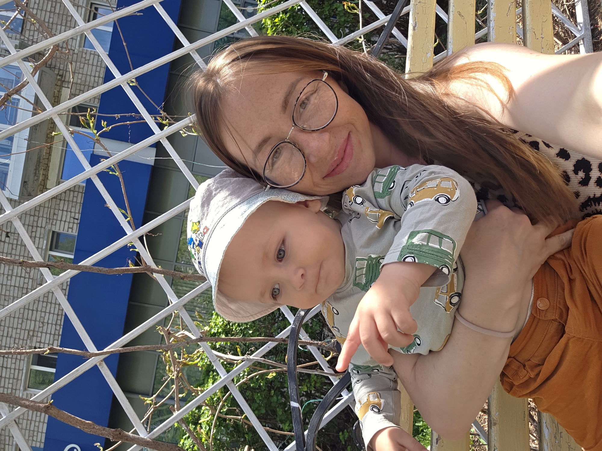 A woman with long brown hair and glasses is holding a baby in her arms. They appear to be outdoors, possibly in a garden or park. The background includes a white trellis-like structure and a building with a blue and white facade. The baby is wearing a hat and a patterned shirt. The woman is wearing an animal-print top and brown pants. The baby is pointing. The location is Chaikovsky, Russia.