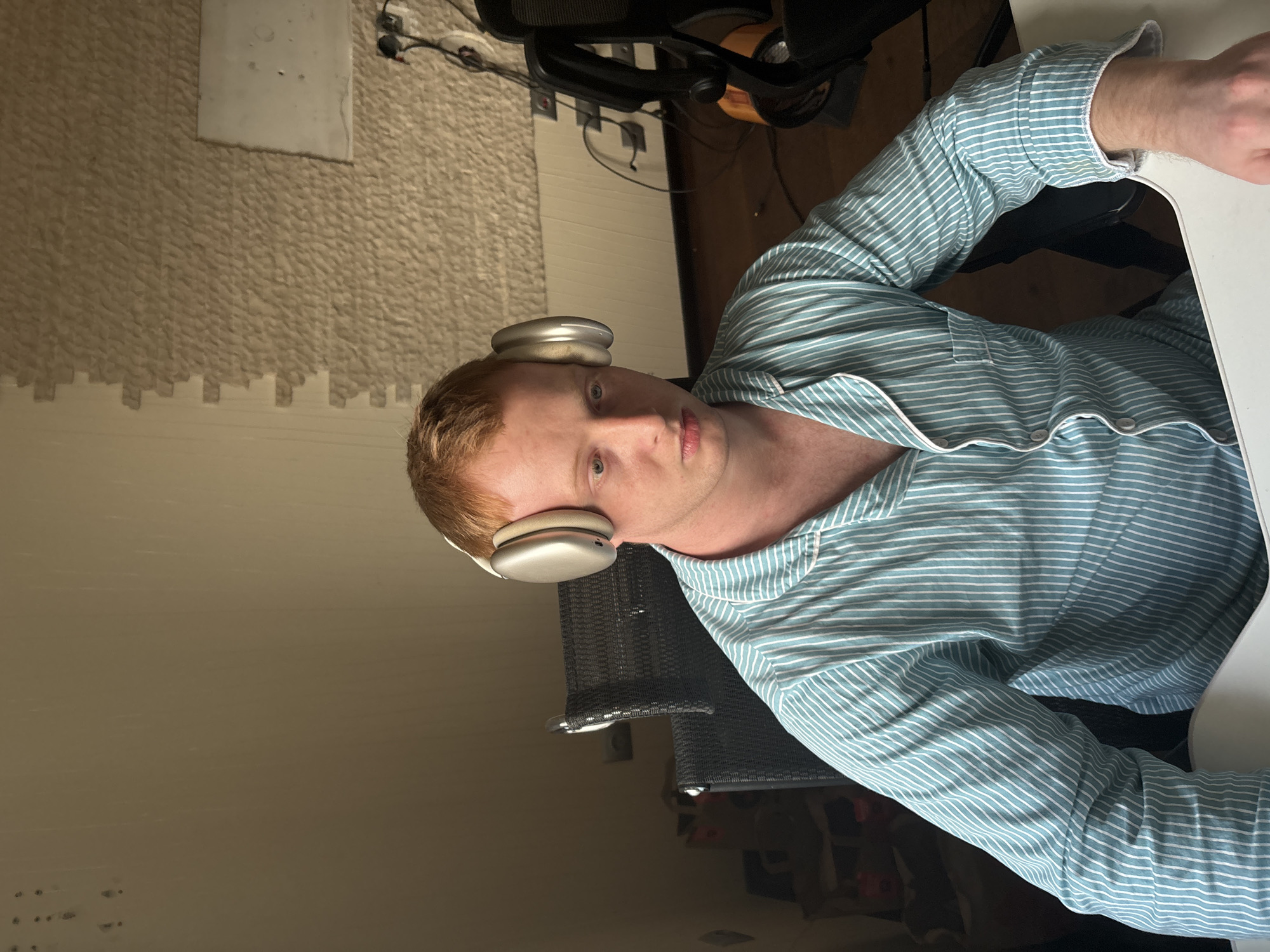 A young man with reddish-blond hair and light blue eyes is depicted from the chest up, looking directly at the camera. He is wearing a light blue and white striped long-sleeved shirt, possibly pajamas or loungewear, detailed with white piping. A pair of silver over-ear headphones rests on his head. His expression is neutral.
He is seated at a white desk, with a portion of his right arm visible on the desk's surface. Behind him, a black office chair is partially visible. The background features a textured wall covering, resembling interlocking foam mats or tiles, with a plain, light-colored rectangular panel affixed to it. Electrical outlets and a cluster of black wires are seen in the upper background. A dark, possibly mesh or fabric, blind or curtain is visible behind his left shoulder. The lighting suggests an indoor setting, likely during the day, with soft, ambient illumination. No discernible text is present. The scene takes place indoors in Ореховка, Russia.