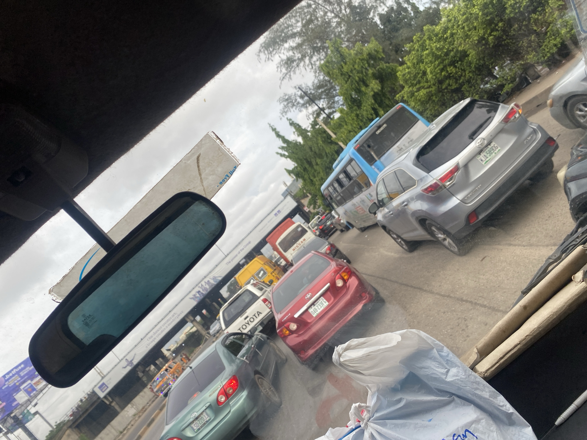 This is a photograph taken from inside a vehicle looking forward, capturing a traffic jam in Ikeja, Nigeria. The scene is dominated by a line of vehicles on a paved road. In the foreground, a large white plastic bag is visible on the left, obscuring a portion of the view. Behind it, several cars are bumper-to-bumper. A green sedan is in the left lane, followed by a red sedan. To the right of these cars, a white Toyota pickup truck is visible, and further back, a yellow minibus and a red and white Nissan bus are also in traffic. A bright blue bus is seen further down the road. The surrounding environment includes trees and buildings in the background, with a cloudy sky overhead. Advertisements are visible on structures above the road, suggesting an urban setting. The overall impression is one of heavy vehicular congestion in a typical Nigerian city environment.