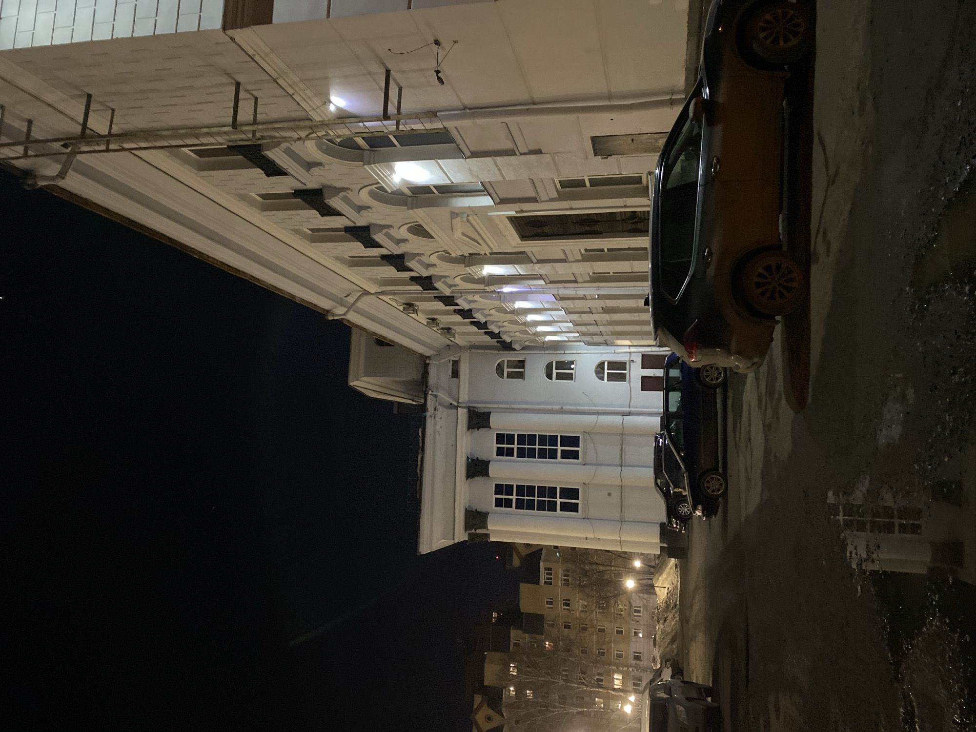 The image captures the Perm Academic Theatre of Opera and Ballet named after Pyotr Ilyich Tchaikovsky, located at Petropavlovskaya St, 25, Perm, Perm Krai, Russia, 614000, during nighttime. The large, multi-story building, rendered in light-colored masonry, dominates the right and center-left of the frame, illuminated by external facade lighting. The building exhibits classical architectural elements, including a prominent corner section with four tall, fluted columns supporting a cornice and tall rectangular windows with white frames. The main facade extends laterally to the right, featuring a series of regularly spaced arched windows, some with dark-colored decorative balustrades or grilles on upper levels, and a large, dark, intricately designed main entrance door. A metal rain gutter and a vertical ladder-like structure are affixed to the right side of the building.
In the foreground and mid-ground, a parking area with an asphalt surface shows wet patches and reflections, indicating dampness or recent precipitation. A dark-colored SUV, heavily soiled with mud on its lower body and wheels, is parked facing left in the foreground-right. Another dark sedan or station wagon is parked further back on the left, near the building's corner. Other vehicles are partially visible on the left. Patches of dirty snow or ice are present on the ground, particularly along the edges of the parking area and near the building. In the distant background-left, multi-story residential buildings with illuminated windows are visible, partially obscured by bare trees, under a dark, clear night sky that displays a single faint star or distant light source. Warm-toned streetlights illuminate the background area. No individuals are present within the scene.