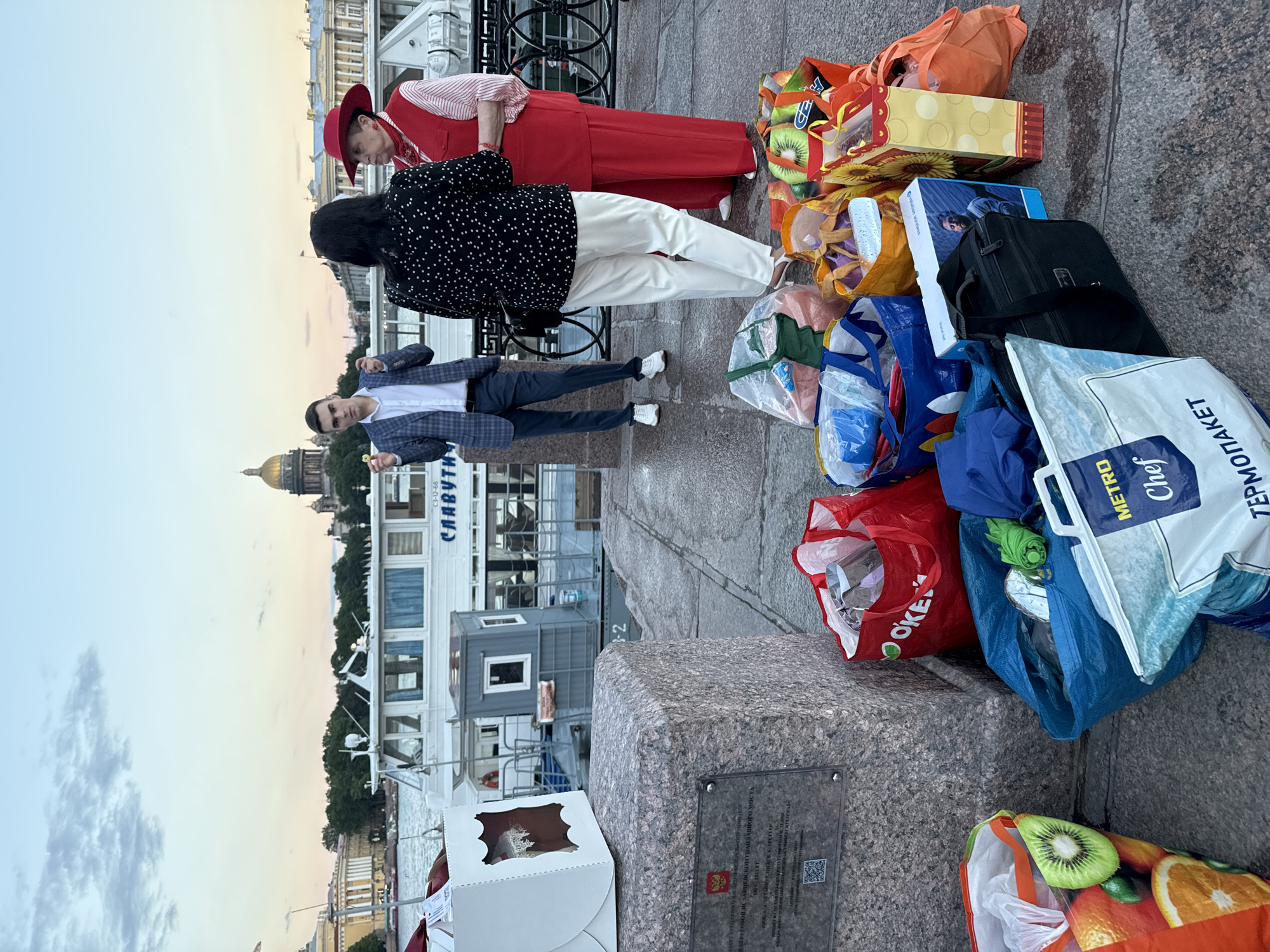 In Saint Petersburg, Russia, on a paved walkway along a river, a group of people stand amidst an assortment of shopping bags. A white boat is docked in the background, with a cityscape featuring a prominent dome visible behind it. The sky is overcast, suggesting either early morning or late evening.
In the foreground, a large pile of shopping bags, some decorated with fruit patterns and others plain, are arranged on the ground. One bag, red with "OKEE" printed on it, stands out. A white box with a lace-like design sits on a stone monument to the left, which bears an inscription and a QR code.
Three individuals are positioned near the bags. A man in a checkered blazer and dark pants stands to the left, holding a small, round object. Two women are to his right. One, wearing a black top with white polka dots and white pants, faces away from the camera. The other, dressed in a red hat, vest, and pants, faces the viewer's left. They appear to be engaged in conversation. The overall scene suggests an outdoor gathering or a stop during an outing, with people and their purchased goods present.