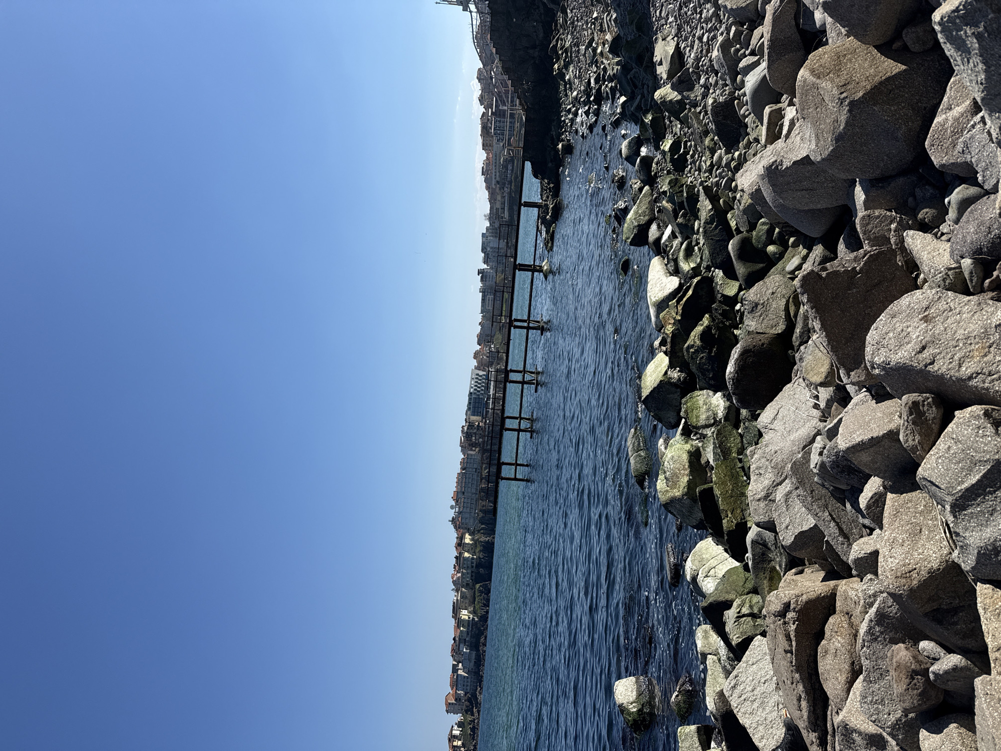 The image shows a waterfront view in Sozopol, Bulgaria. A rocky shoreline transitions into the sea. A pier extends out into the water, with buildings visible along the coastline in the background. The sky is clear and blue. The rocks are large and varied in color, with some showing signs of moisture or algae. No people are visible in the shot.