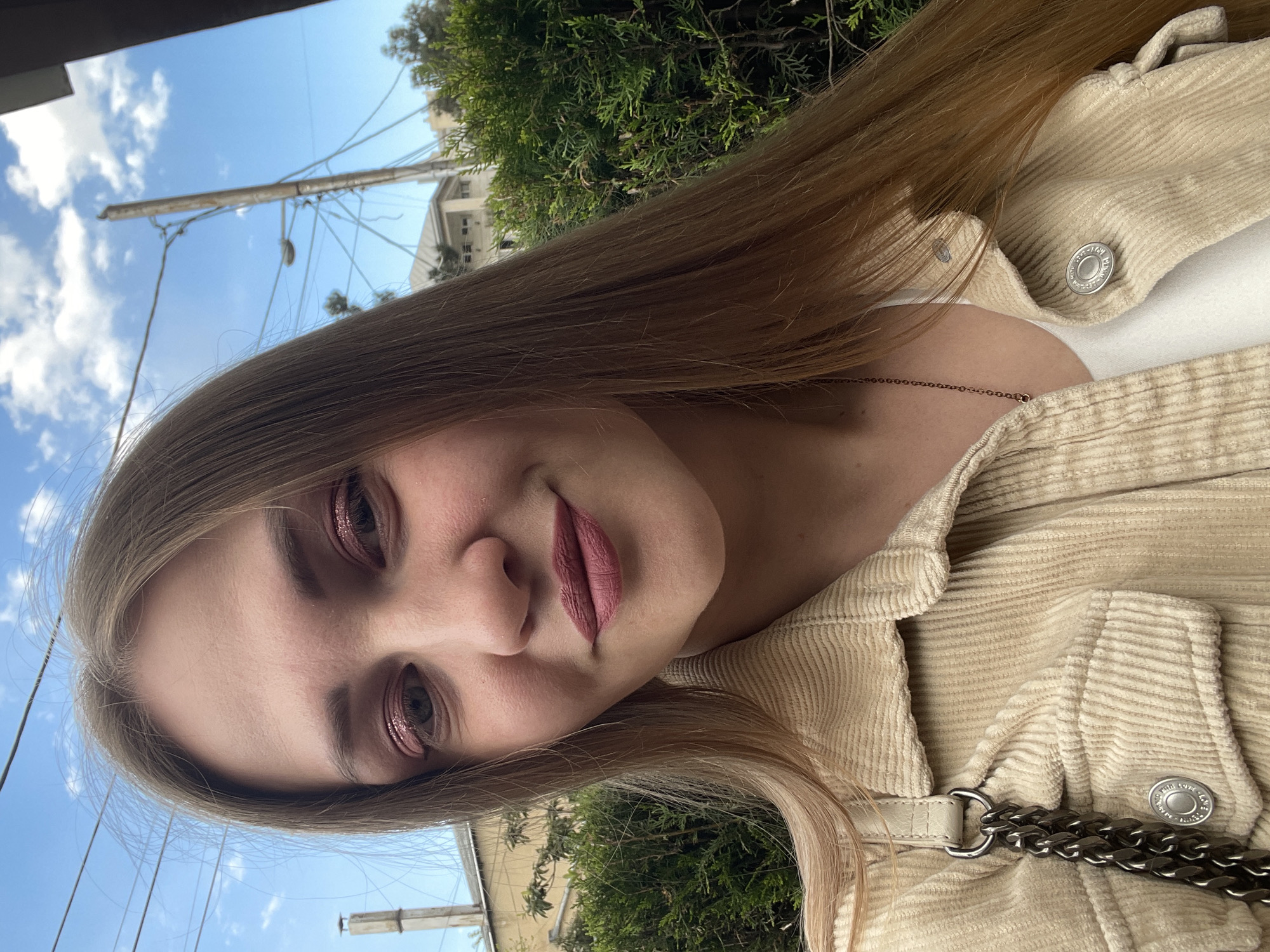 A woman is outdoors in what appears to be Tbilisi, Georgia. She's wearing a tan corduroy jacket over a white top, with a silver chain detail visible. She has long blonde hair and is wearing rose-colored eyeshadow and lipstick. The background shows trees, buildings, a blue sky with clouds, and power lines.