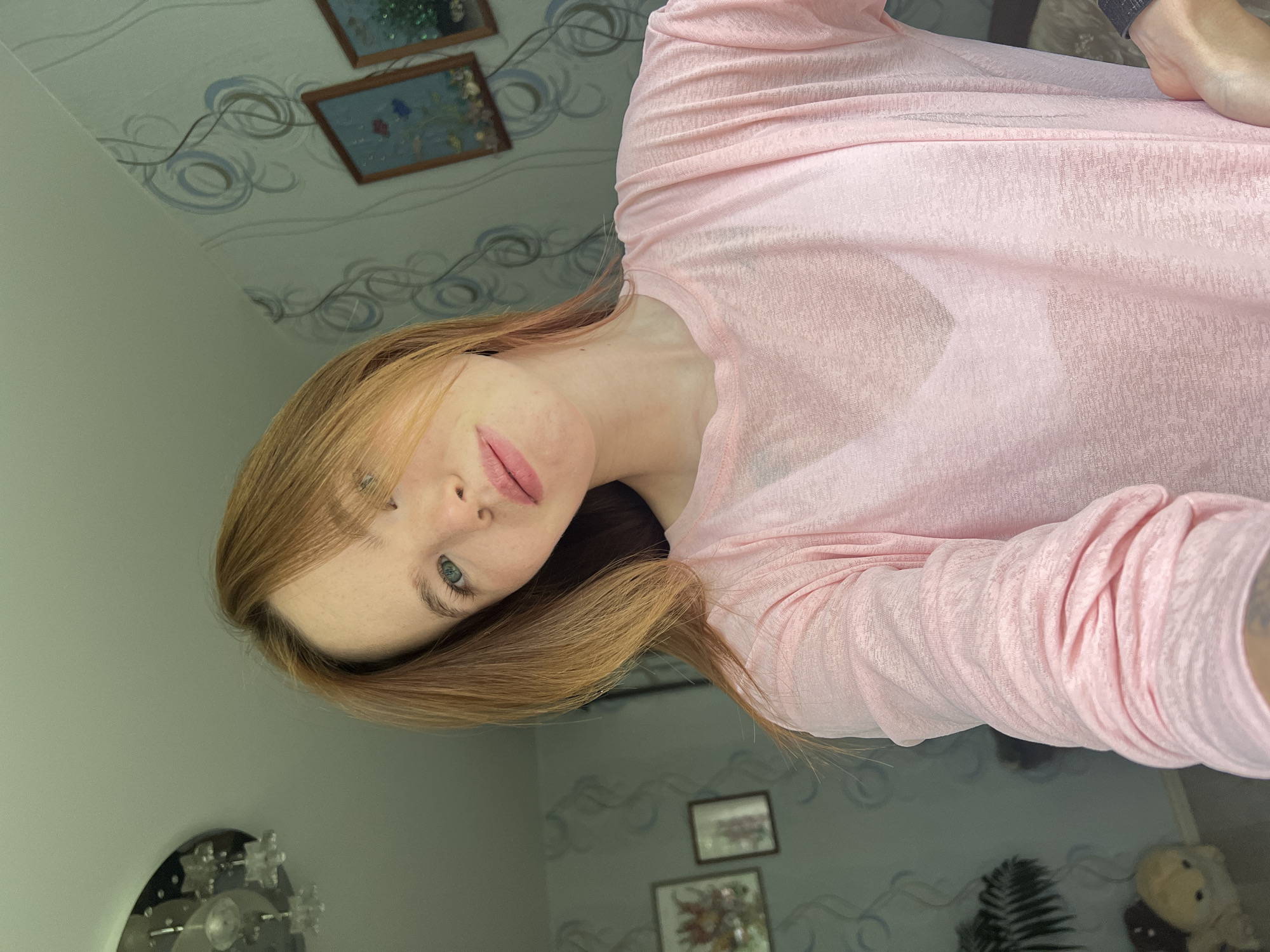 A selfie shows a woman with red-blond hair and blue eyes wearing a pink shirt, taken from an upward angle. The location appears to be indoors, potentially in a home in Chaikovsky, Russia, given the presence of decorative paintings on the wall and objects like a chandelier and teddy bear in the background. The photo focuses primarily on the woman's face and upper torso.