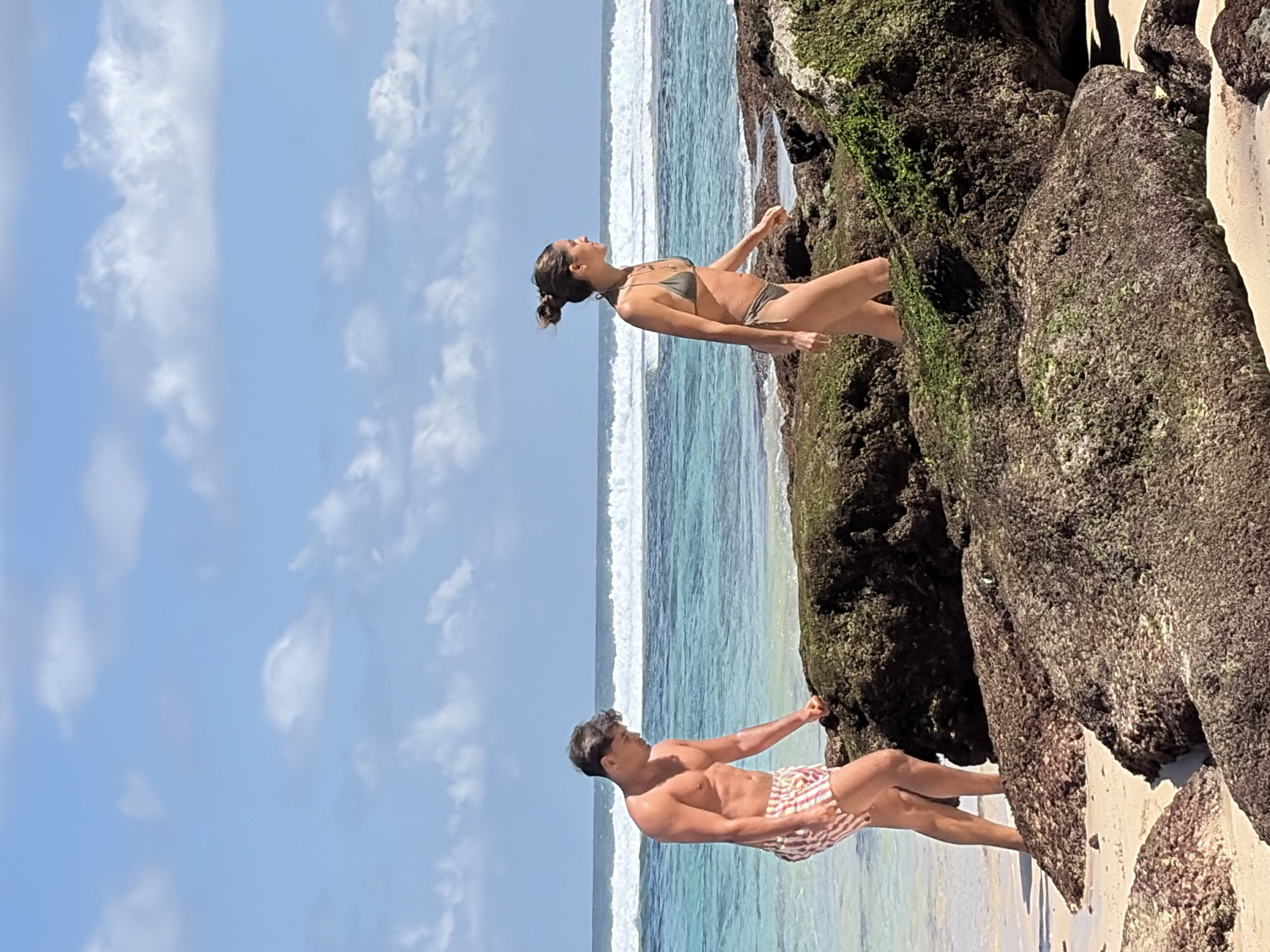 A man and a woman are walking on a rocky beach in Kuta Selatan, Indonesia. The man is wearing striped swim trunks and is looking down at the rocks. The woman is wearing a bikini and is walking away from the camera, looking out at the ocean. The ocean is turquoise and there are waves breaking in the distance. The sky is blue with scattered clouds. The rocks are covered in green moss. The sand is light brown.