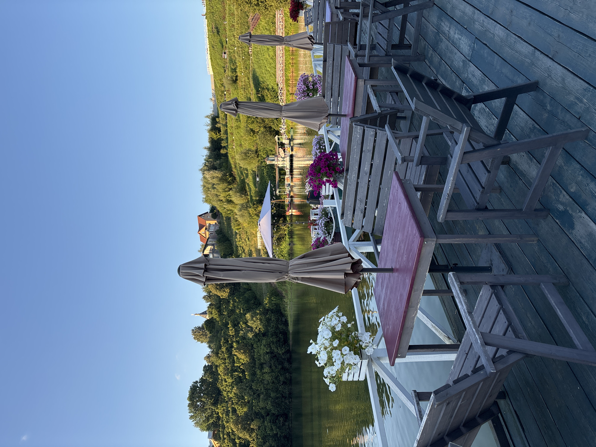 The image depicts an outdoor seating area on a wooden deck next to a body of water. The deck is furnished with several wooden tables with reddish-brown tops and matching wooden chairs and benches. Umbrellas, some folded and some open, are positioned near the tables. Potted flowers, including white and pink varieties, are placed along a white railing that separates the deck from the water. In the background, a green, tree-covered hillside rises, with houses visible at the top. A small structure and a bridge are situated on the water. The sky is clear and blue. The scene is likely a restaurant or cafe located by a lake or river, possibly in the village of Romashkovo, Russia, given the architectural style and natural landscape. There are no people visible in the image.