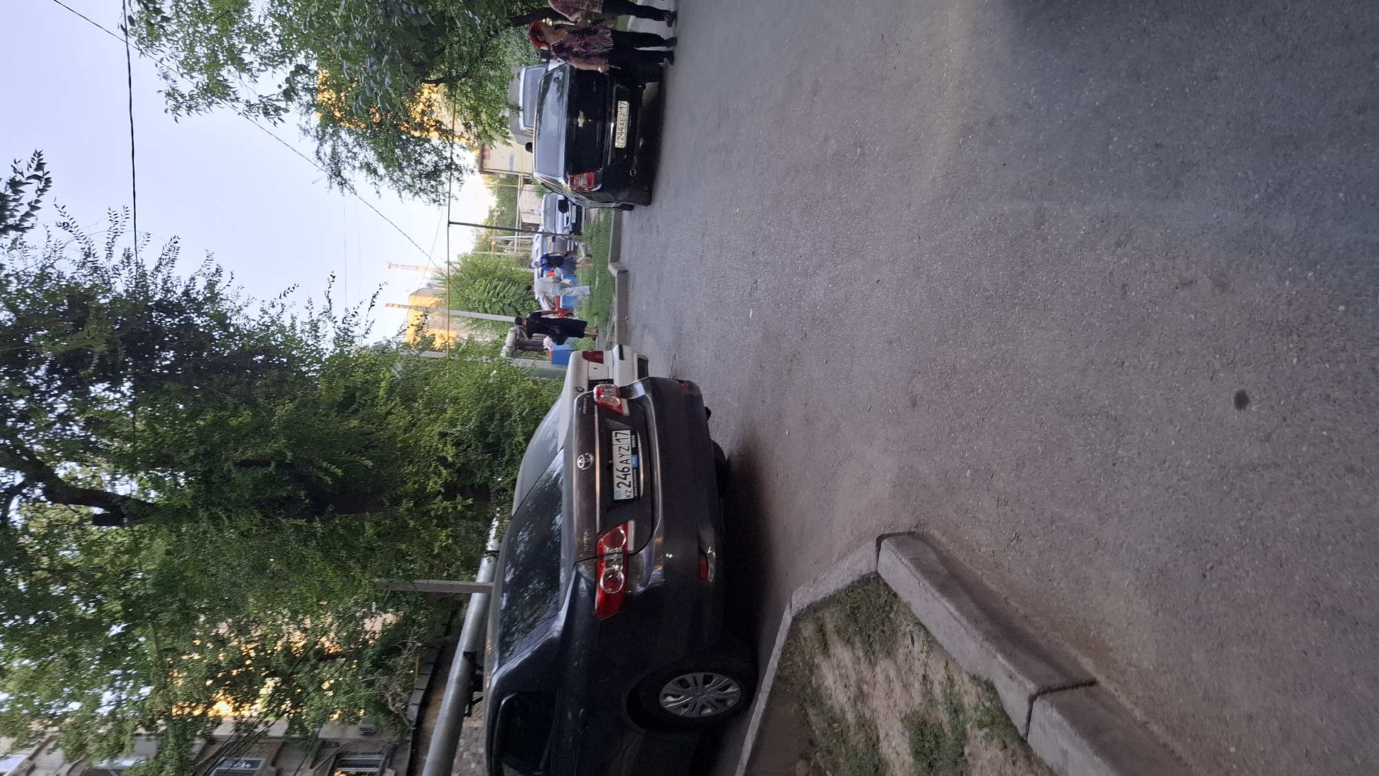 A group of people are standing by parked cars on a street in Shymkent, Kazakhstan. The scene appears to be outdoors, with trees and buildings visible in the background. The sky is clear, suggesting it is daytime. The people are dressed in casual clothing, and their body language does not suggest any particular emotion. The cars are parked along the side of the road, and some of them have license plates visible. One car has the license plate "246 AYZ 17". Another car has the license plate "E 244 AB 17". The overall impression is of a normal street scene, possibly in a residential area. The lighting suggests it might be late afternoon or early evening due to the warm tones filtering through the trees.