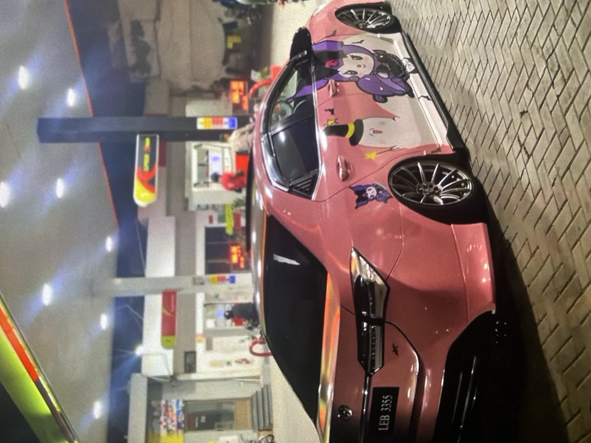 A pink car with anime-style graphics on the side panels is parked at a gas station. The license plate number visible on the car is "LEB 3355." The location is Lhokseumawe, Indonesia, as suggested by the surroundings and the license plate format. The gas station has yellow and red branded signage. People are visible inside the gas station store. The ground is paved with brick tiles.