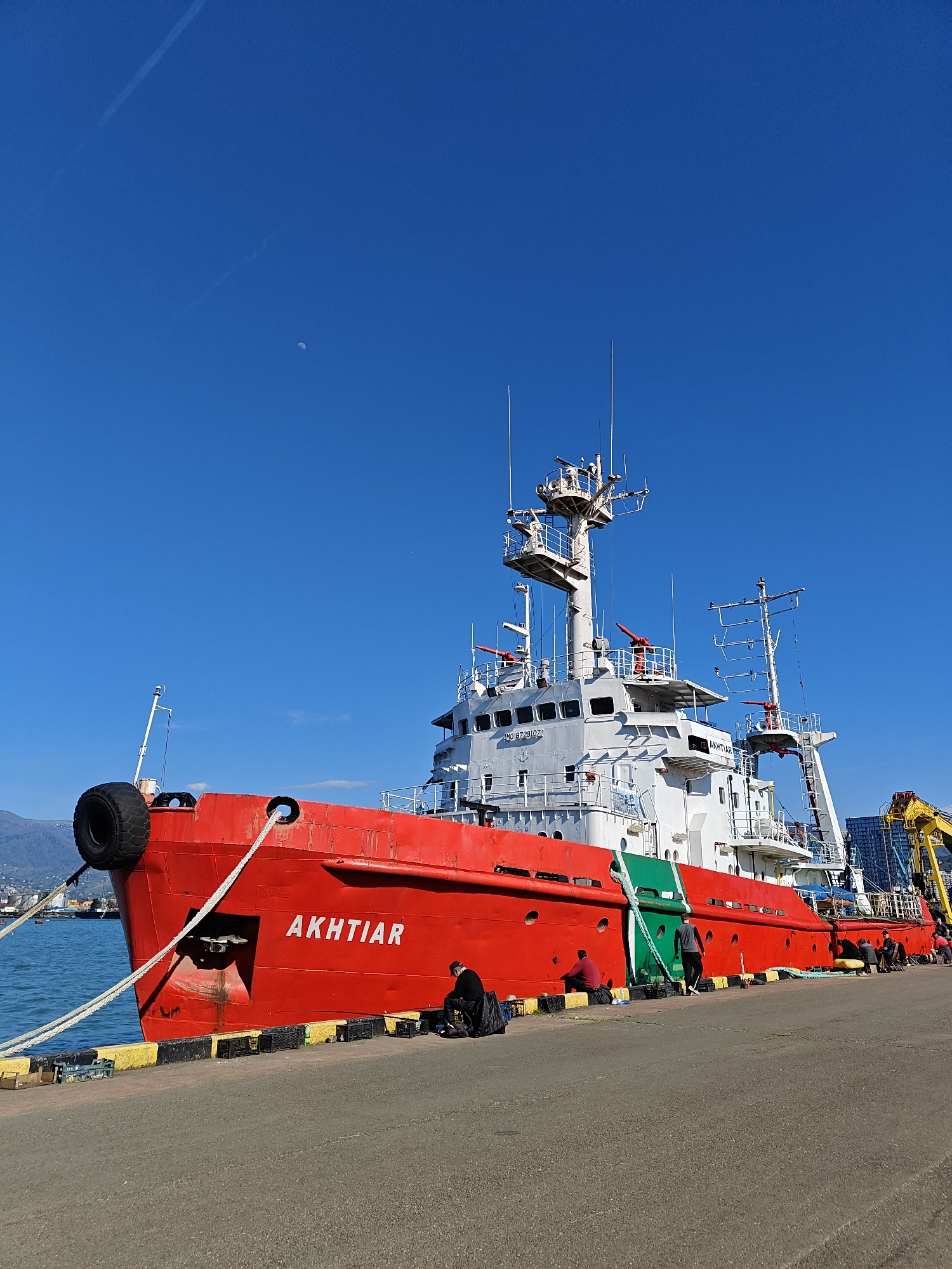 The image shows a red and white ship named "AKHTIAR" docked at the port in Batumi, Georgia. The ship is a large vessel with multiple decks, antennas, and a crane visible on the rear. It is positioned alongside a concrete pier with black and yellow painted edges. Several people are seen sitting or standing near the ship's hull on the pier. In the background, there are mountains and other port facilities visible across the water. The sky is clear and blue, with a faint crescent moon visible in the upper left. The atmosphere appears to be calm and sunny.