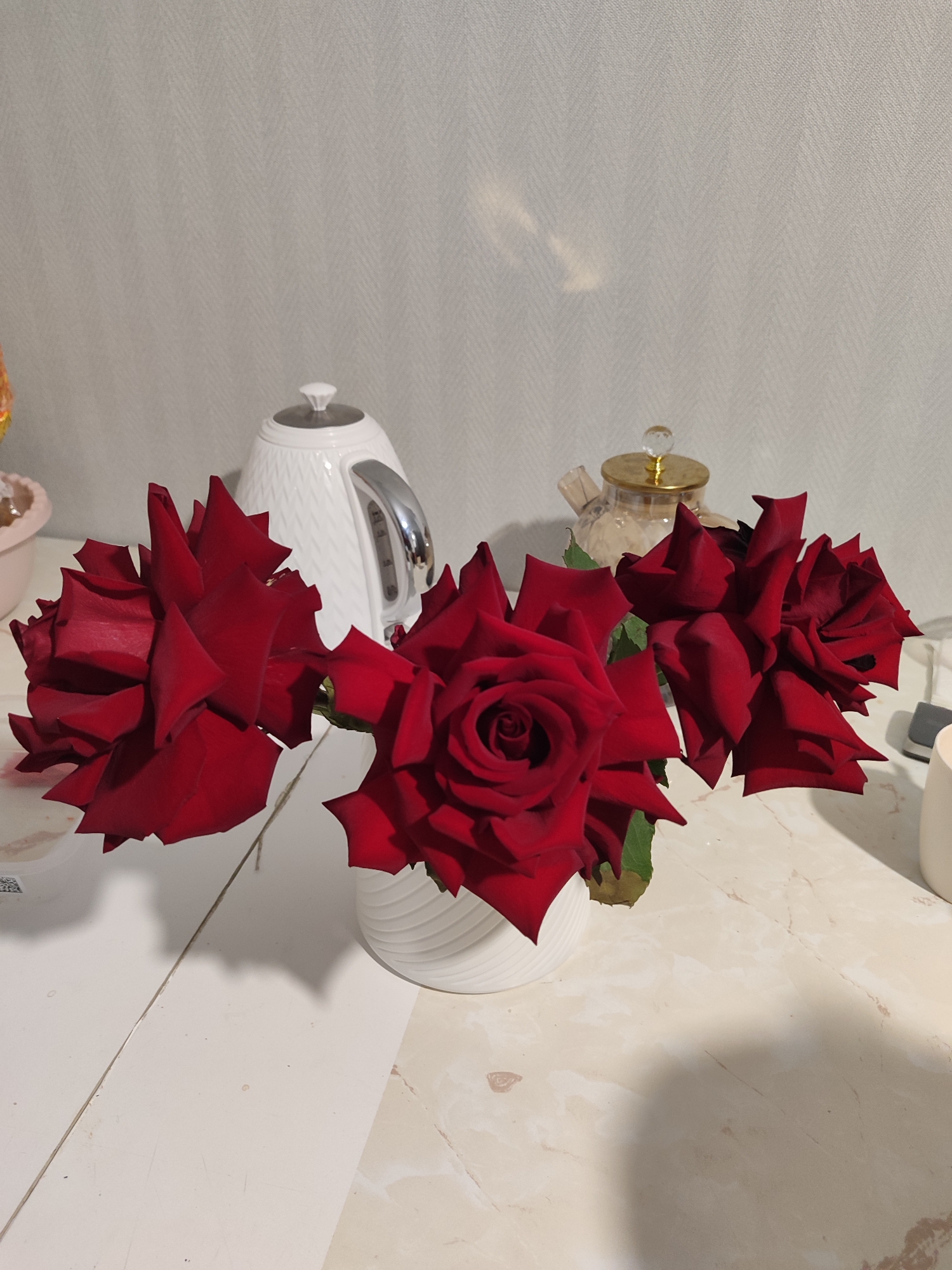 The image displays a still life composition featuring three vivid red roses arranged in a white, ribbed vase. The roses, with their deep crimson petals and subtle green foliage, are positioned centrally on a tabletop.
The setting appears to be an indoor space, possibly a kitchen or dining area. In the background, to the left of the roses, a white electric kettle with a patterned, possibly diamond-textured, surface and a metallic handle is visible. To the right, a small, clear glass teapot with a golden lid and a clear, faceted knob is partially seen. The background wall features a light grey or off-white hue with a distinct herringbone or chevron pattern.
The tabletop itself is comprised of two distinct sections: the left side is a plain, light-colored surface, possibly wood or laminate, with a visible seam running horizontally, while the right side exhibits a light beige or cream-colored marble pattern. Soft shadows are cast by the roses and other objects, indicating a well-lit environment, likely illuminated by indoor lighting or daylight.
No specific activity is taking place; the scene is a static arrangement of decorative items. There are no people visible, and no discernible text is present on any objects. The overall impression is one of domestic tranquility and aesthetic arrangement. No specific visual cues link the scene to Izhevsk, Russia.