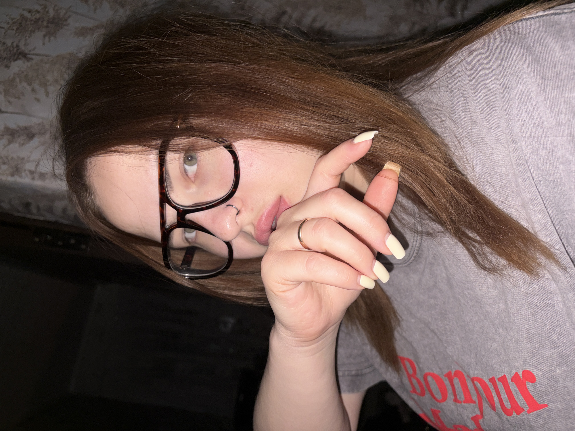 A woman with shoulder-length brown hair, wearing glasses, is oriented diagonally to the left in the frame. She has a nose ring and is wearing a gold ring on her finger. Her fingernails are painted a light yellow color. She is wearing a gray t-shirt with the word "Bonjour" in red lettering visible. The background is dark, but a bed with a patterned headboard is faintly visible in the upper left corner. There is no information available to indicate a specific location in Port-Katon, Russia.
