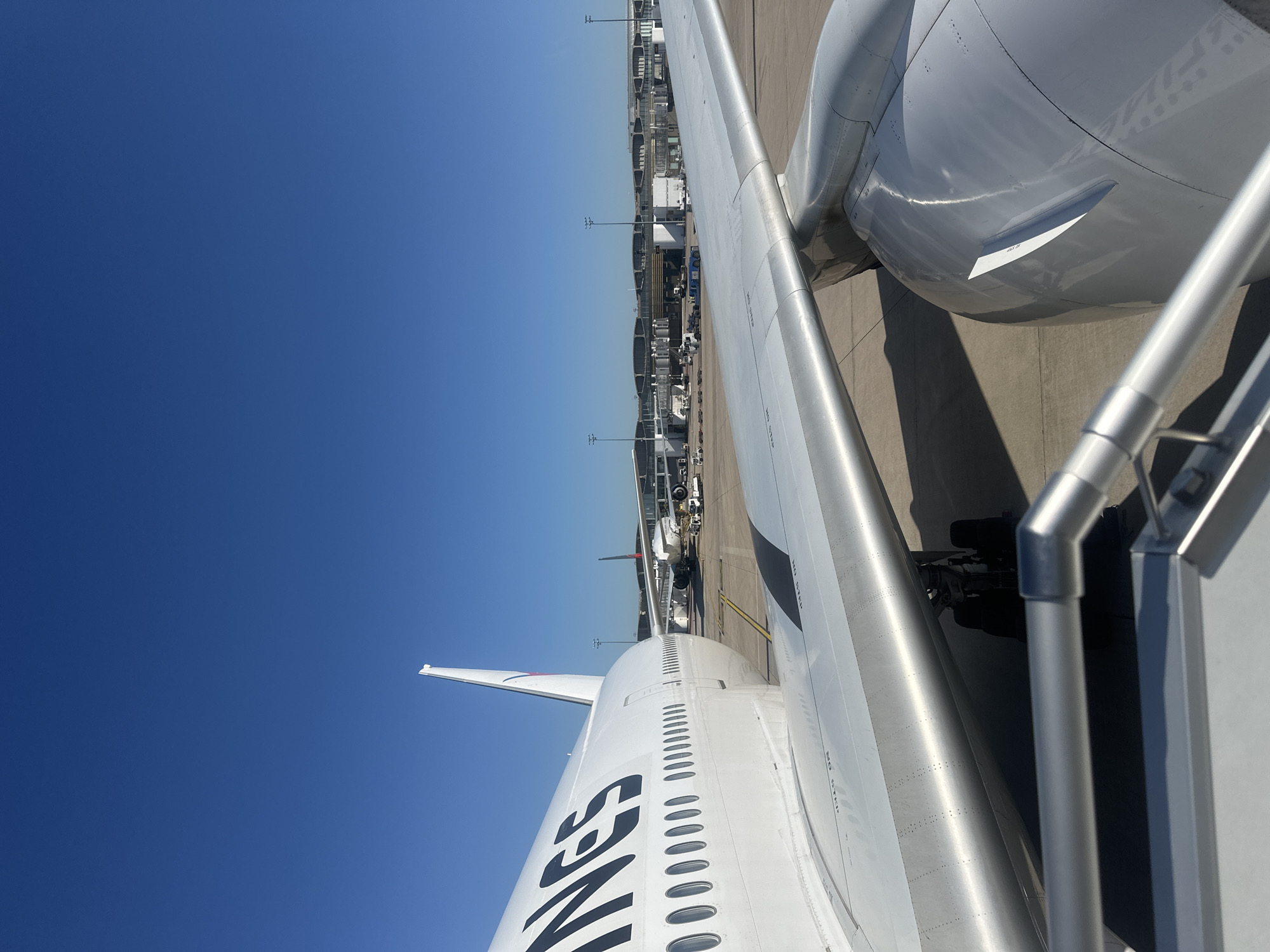 This image depicts an airplane at Charles de Gaulle Airport, located in Tremblay-en-France, France. The viewpoint is from a passenger boarding bridge, looking onto the wing and engine of the aircraft. Visible markings on the fuselage read "NAS". Another aircraft is visible further down the tarmac alongside airport terminal infrastructure. The sky is a clear blue.
