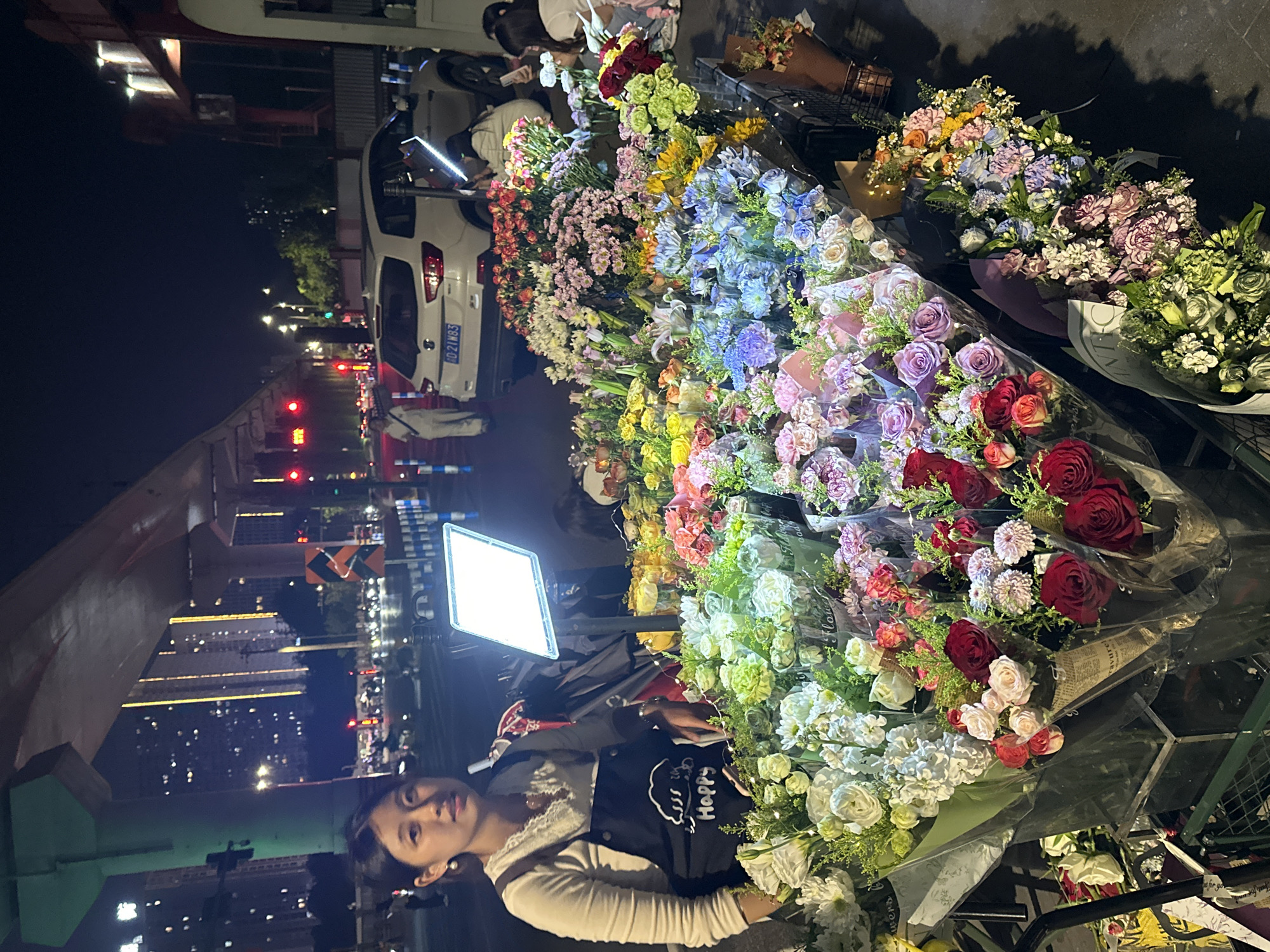 The image shows a woman standing in front of a large display of flowers, likely a flower stand or stall, in an urban environment at night. She has long dark hair and is wearing a white sweater over a black apron with a doodle and the word "Happy" on it. The flower stall is filled with various kinds of colorful flowers wrapped in plastic, including roses, lilies, and other blooms. The background shows a city street with buildings, streetlights, and car headlights. A white car is visible behind the flower stall. Based on the license plate number "D21N83" and other visual clues, the location is the Chongqing High-tech Zone, China.
