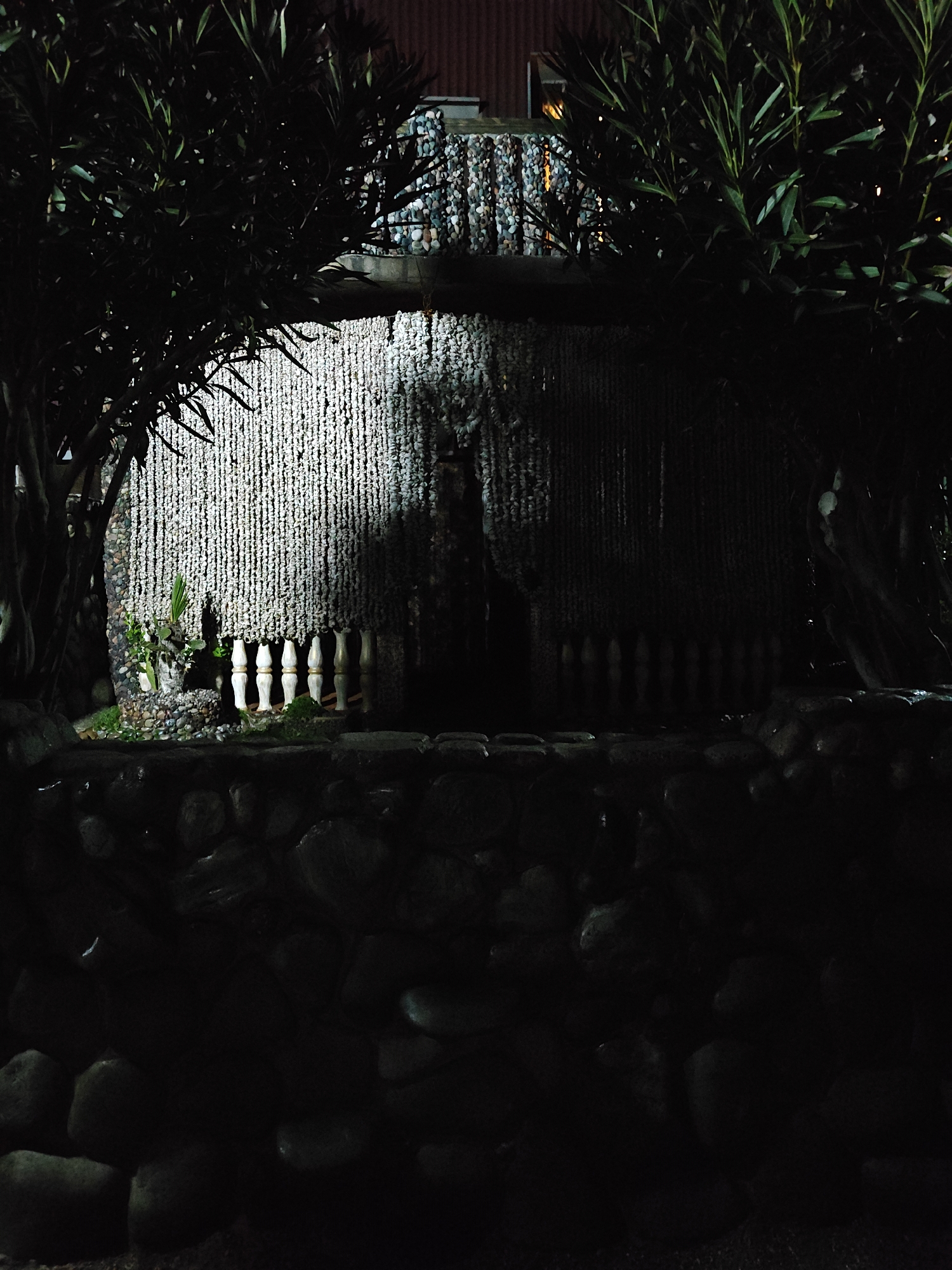 The image depicts a dimly lit scene, likely at night, featuring a structure adorned with hanging strings of what appears to be shells or beads, creating a curtain-like effect. The structure is framed by dense foliage on either side, suggesting a garden or outdoor setting. In front of the "curtain," there is a small decorative fence made of white posts, with some greenery visible behind it. The foreground is dominated by a wall constructed of large, dark stones. The overall atmosphere is mysterious and somewhat obscured due to the low lighting conditions. The location is stated as Unknown, Unknown.