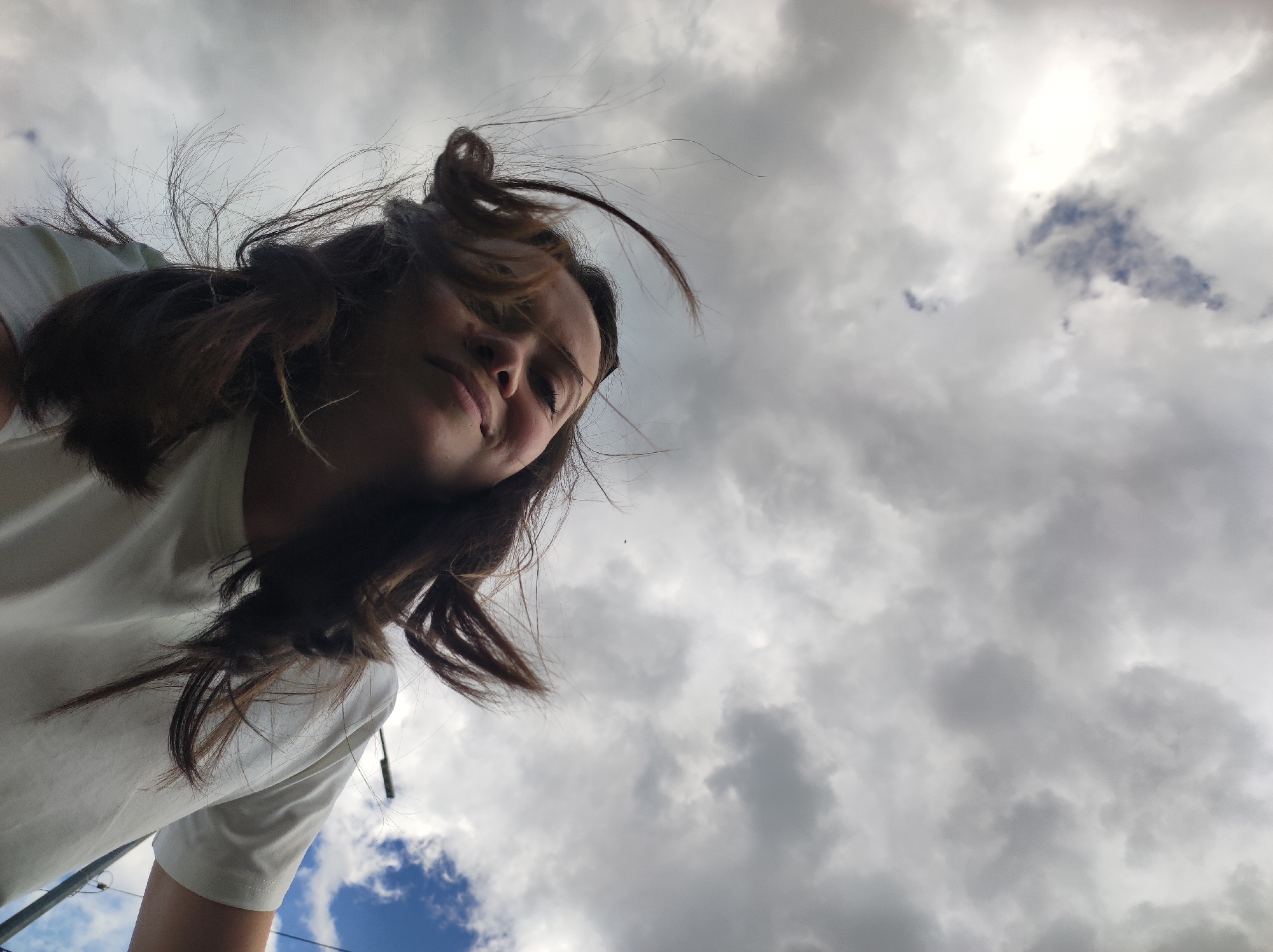 A low-angle shot shows a person with medium-length brown hair standing outdoors with a cloudy sky above. The person is wearing a white shirt. The hair is tousled by the wind. Part of a utility pole is visible at the bottom left corner of the frame. The location is Chaikovsky, Russia.