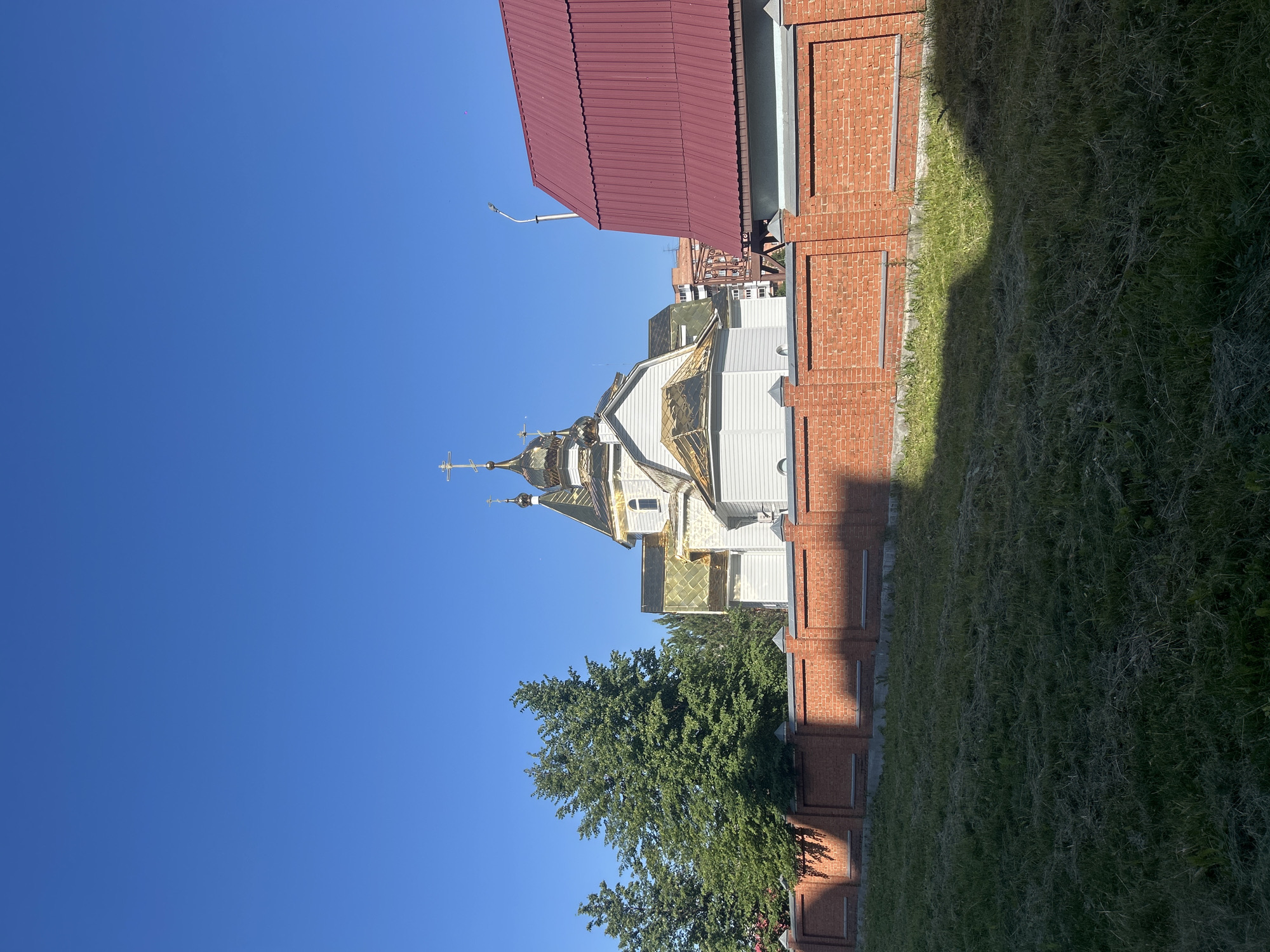 The image shows a church with golden domes and a white facade, situated behind a red brick fence. Adjacent to the church is a building with a red metal roof. In the foreground, there is a grassy area. The sky is clear and blue. The scene appears to be located in the city of Kemerovo, Russia, based on the presence of typical architecture and landscaping for the region. There are no people visible in the image.
