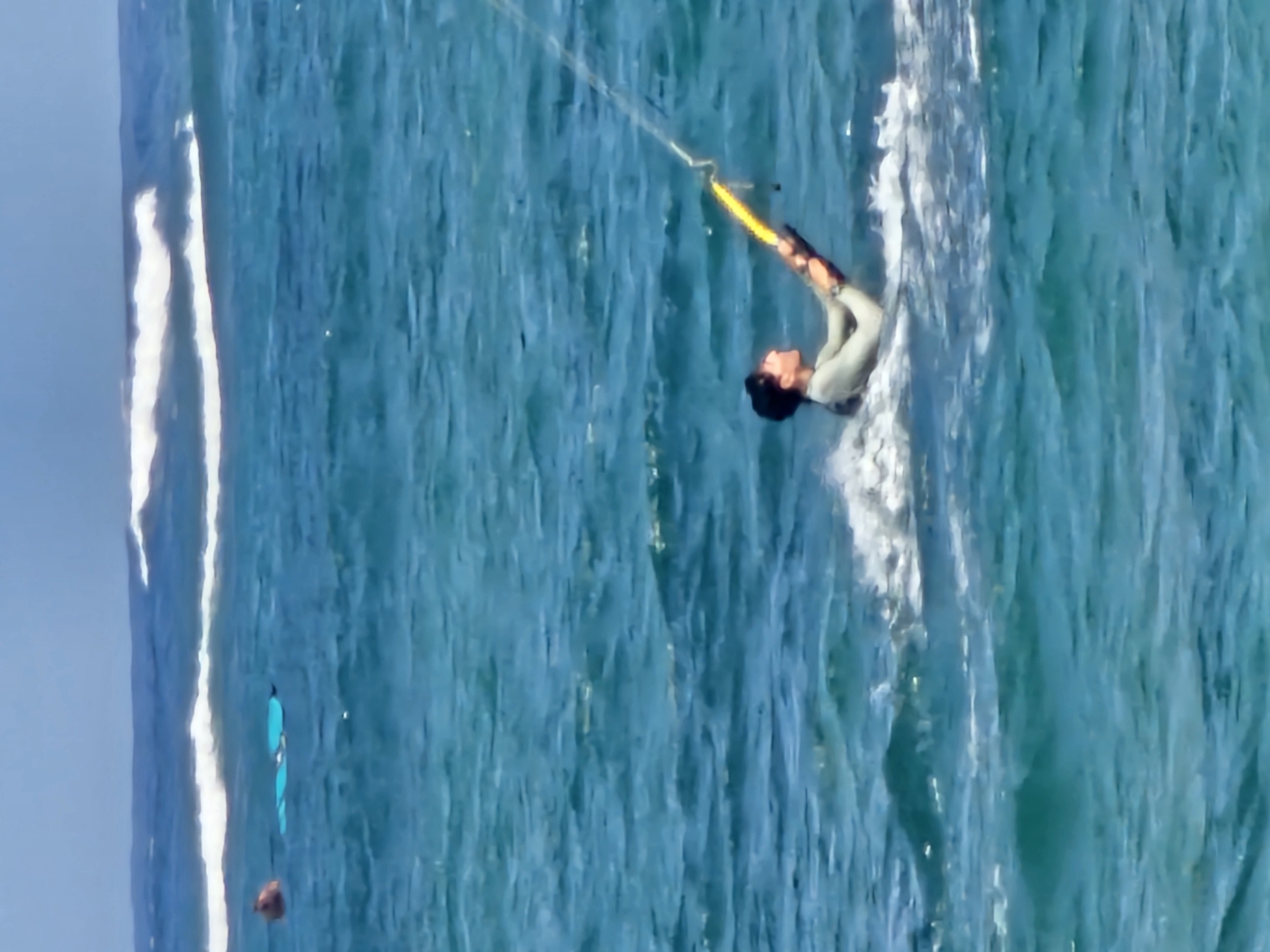 A person is kitesurfing in the ocean. The person is wearing a light-colored wetsuit and is holding onto a yellow kite bar with both hands. The kitesurfer is leaning back, being pulled through the water, creating a white wake. In the background, there are two blue boards floating in the water and a person's head visible above the surface. The ocean water is a deep blue with some white wave crests in the distance. The sky is a light blue with some clouds. The image was taken on a sunny day, likely at a beach in Denpasar, Indonesia, known for its watersports.