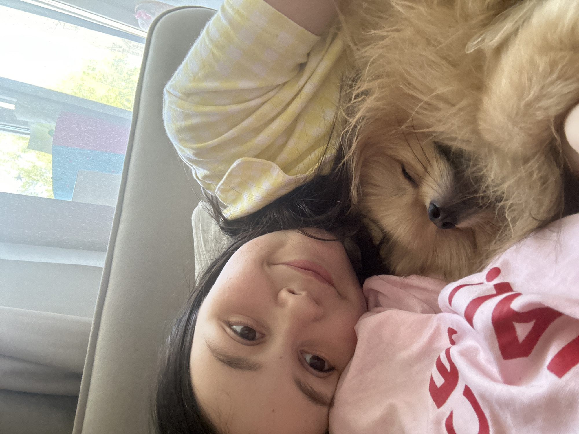 A young girl with dark hair and brown eyes is lying down, with a fluffy, tan dog nestled against her. She is wearing a yellow checkered shirt on one side and a pink shirt with red lettering on the other. They appear to be on a light-colored couch near a window. The scene is not identifiable to the city of Votkinsk, Russia.