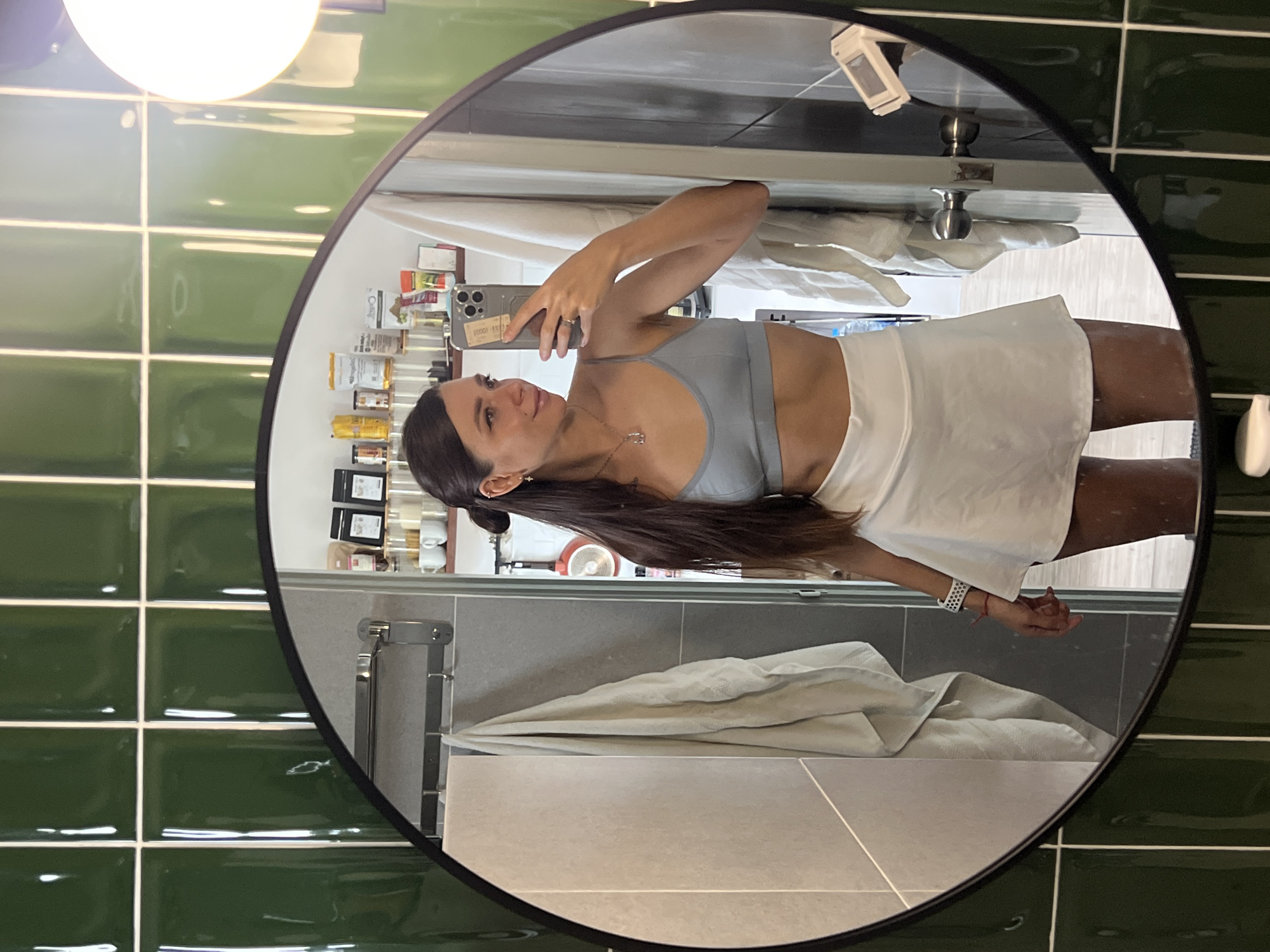A mirror in an Unknown, Unknown bathroom reflects a person taking a selfie with a mobile phone. The person is wearing a light blue sports bra and a white skirt. The bathroom has green tiled walls, a gray wall with a towel rack, and a white towel. There are various items on a shelf, including containers and a white object. A light fixture is visible in the upper left corner.
