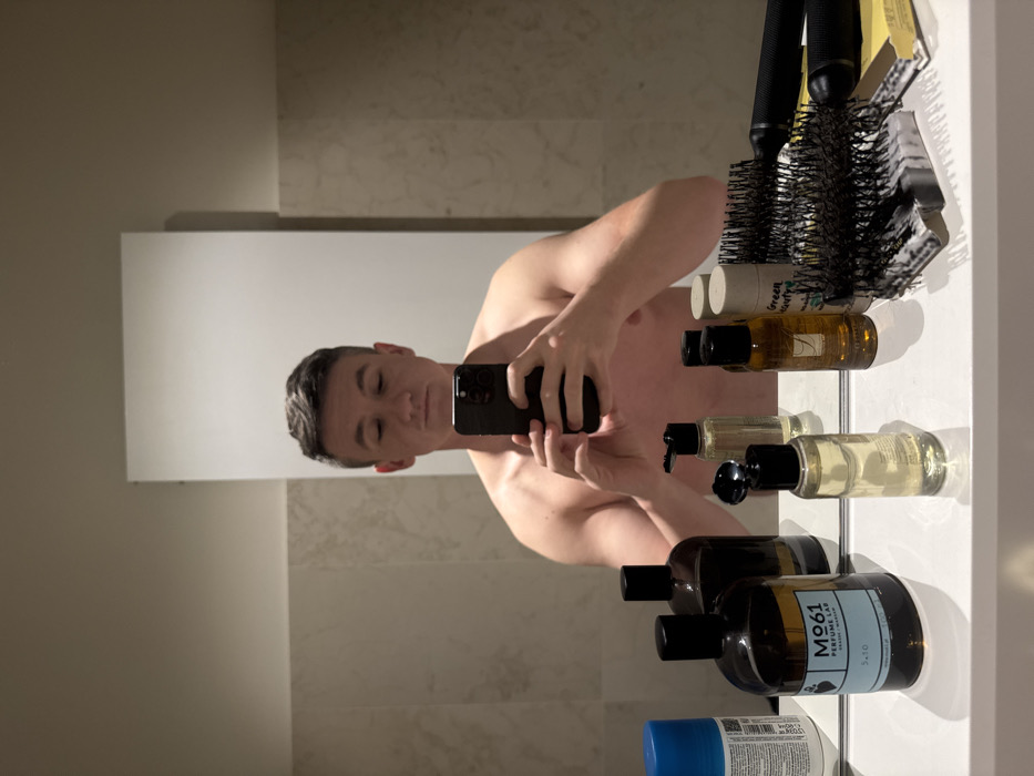 A shirtless adult male is taking a mirror selfie inside a bathroom in Venice, Italy. The man, with short, dark, styled hair, holds a black smartphone horizontally with both hands, positioned to photograph his own reflection. His eyes appear closed or are directed downwards. The white countertop in the foreground displays a collection of personal care items. These include a large dark amber bottle labeled "MØ61 PERFUME LAB," a smaller amber bottle labeled "ACQUA DI PARMA," and a cylindrical white container marked "Green Beauty." Several other bottles, including clear ones with black pump and flip-top dispensers, are also present, alongside a blue aerosol can, two large round hairbrushes (one black and one with a metallic barrel), and a smaller rectangular brush. The bathroom features light-colored, possibly beige, tiled walls, and the mirror reflects a plain white vertical panel or door behind the man.