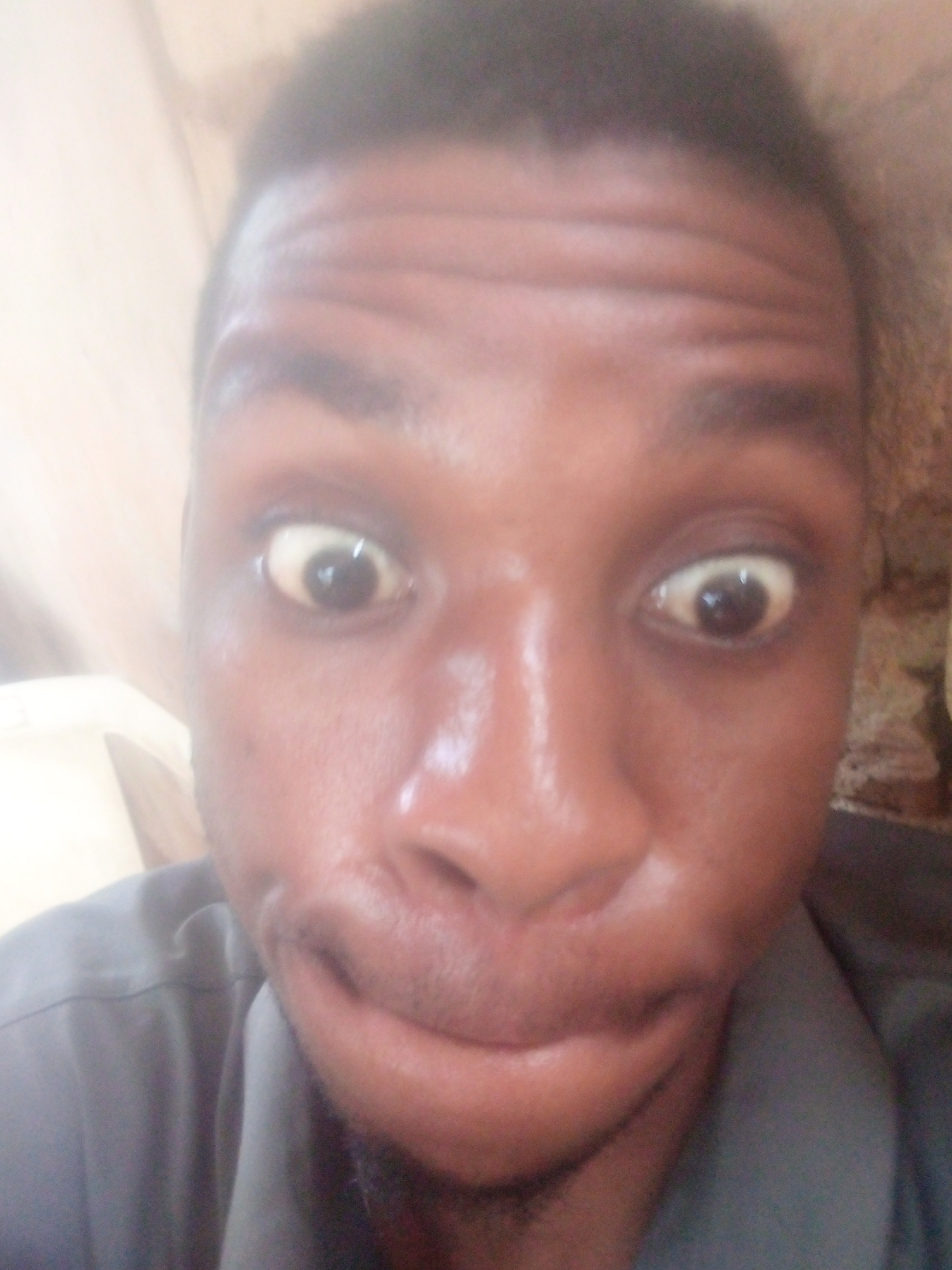 A close-up, low-resolution portrait image features an adult person with dark skin and short, dark hair, likely a selfie. The person is centered in the frame, looking directly at the camera with a highly exaggerated expression. Both eyes are wide open, with the sclera appearing yellowish, and the pupils are dark. The eyebrows are raised, creating prominent horizontal wrinkles across the forehead. The mouth is distorted, possibly in a pout or grimace, with the lips pushed forward and a slight downward curve. Visible stubble or very short beard growth is present on the upper lip and chin area.
The person is wearing a dark grey or blue collared shirt, visible across the shoulders and chest. The background is out of focus and indistinct, suggesting an indoor setting. On the left side, the background appears light beige or cream, while the right side shows a darker, textured brown surface, possibly a wall. The lighting is soft and appears to be from the front, creating some highlights on the nose and forehead. No text is visible in the image.