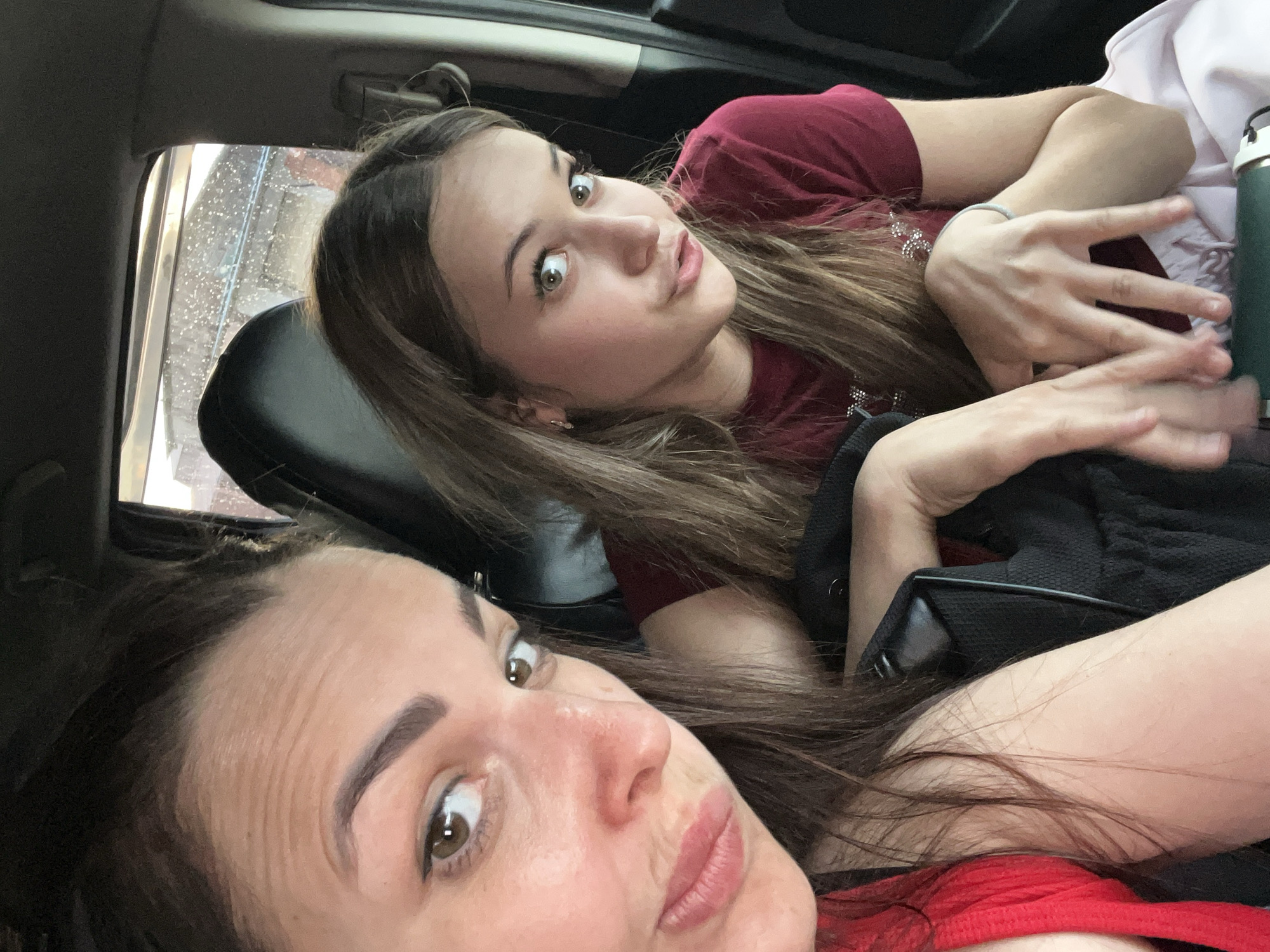 This is a selfie taken from inside a vehicle. Two young women are the subjects of the image. The woman in the foreground is positioned lower and to the left of the frame, with her head tilted back, and her face partially visible. She has dark brown hair and brown eyes, and is wearing a red top. The second woman is in the background, to the right, lying down and looking at the camera. She has long, light brown hair, light-colored eyes, and is wearing a maroon shirt. Her arms are crossed over her chest, and she is wearing a bracelet on her left wrist. A green thermos is visible behind her. The interior of the car is dark, with the exception of the light coming through the windows, suggesting it might be daytime. The mood appears casual and relaxed, likely a candid moment captured during a car ride. There is no visible text in the image.