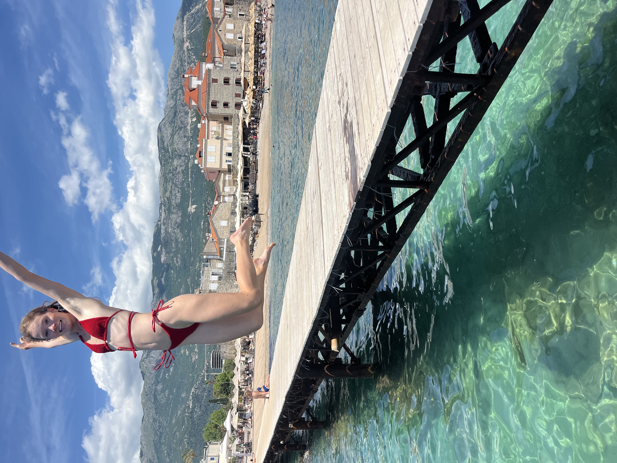 A young woman with reddish-brown hair, wearing a red two-piece bikini, is captured mid-air, jumping from a wooden pier into the clear, turquoise-blue water in Budva, Montenegro. Her arms are extended upwards, and her legs are bent in a jumping motion, with a smile on her face. The wooden pier has dark, rusty metal support structures visible beneath it. The water is highly transparent, revealing the seabed and reflections. In the background, a sandy beach is visible, densely populated with white umbrellas and lounge chairs, where numerous people are relaxing. Several traditional stone buildings with distinctive reddish-orange tiled roofs characterize the coastal town behind the beach. Looming in the far background are large, rugged mountains under a blue sky with scattered white clouds. One person in a blue swimsuit is distinctly visible sitting on the beach near the pier's end.