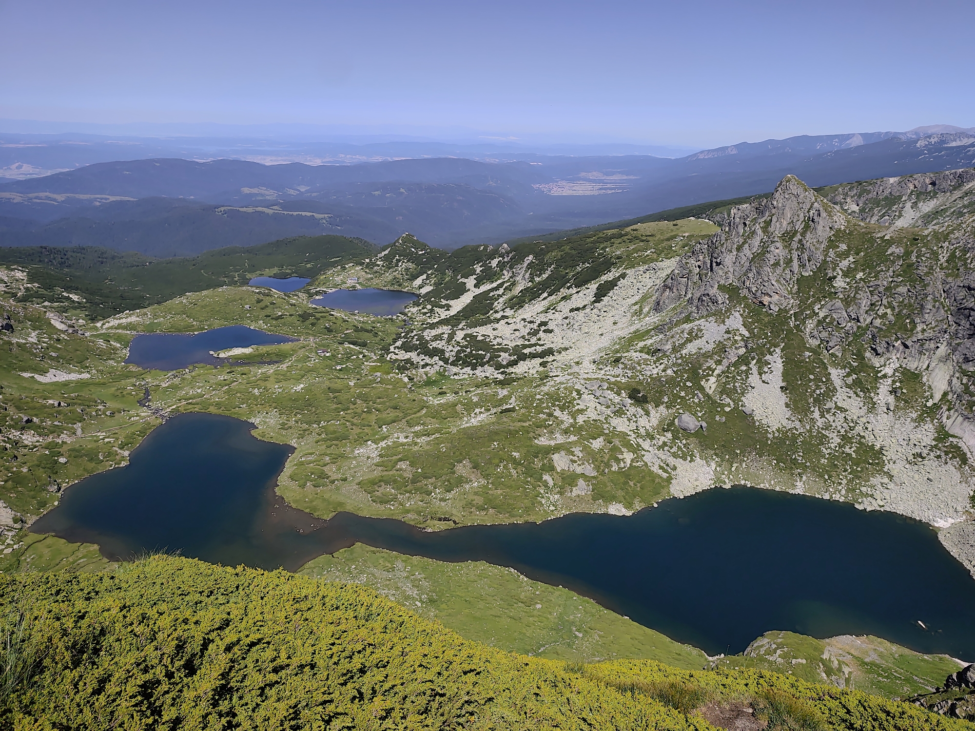 This image captures a panoramic view of a mountainous landscape with multiple glacial lakes. The foreground is dominated by a dense, low-lying green shrubbery with small yellow flowers, suggestive of alpine vegetation. Beyond this, a series of dark blue lakes are visible, nestled within the undulating terrain. These lakes vary in size and shape, with some appearing connected by narrow water channels. The surrounding slopes are covered in a patchwork of green grass, sparse rocks, and larger scree fields. In the mid-ground, jagged rocky peaks rise, some with exposed stone faces. Further in the distance, successive ranges of hazy blue mountains recede towards the horizon under a clear, pale blue sky. The scene is devoid of any human presence or man-made structures, indicating a remote, natural environment. This specific location is known as the Seven Rila Lakes in Rila National Park, Bulgaria, which is near the Rila Monastery (Rilski manastir).