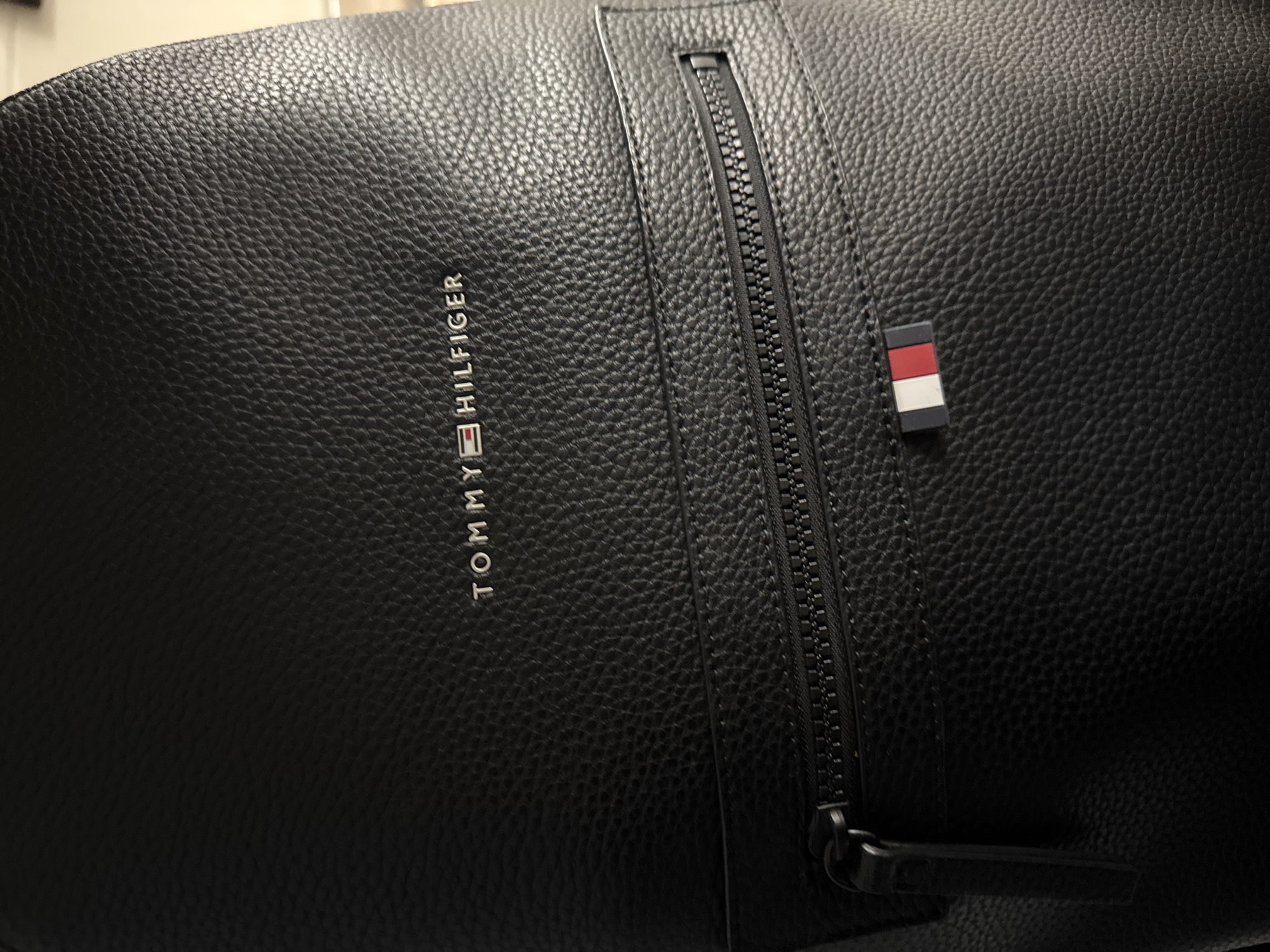 A close-up shot of a black Tommy Hilfiger bag. The bag is made of a textured material, possibly leather or a leather-like synthetic. The Tommy Hilfiger logo is vertically oriented on the left side of the frame. A black zipper is visible next to the logo, with a zipper pull at the bottom. A small Tommy Hilfiger flag logo is positioned to the right of the zipper.