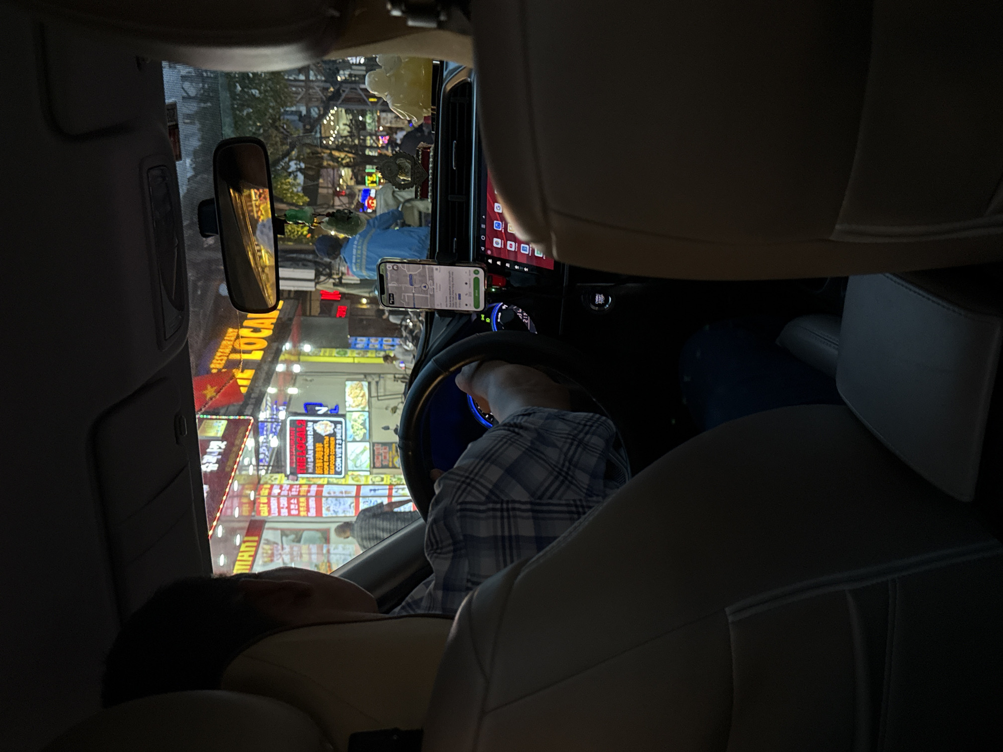 The image depicts the interior of a car in Nha Trang Ward, Vietnam. A man is reclined in the driver's seat. A cell phone attached to a dashboard mount displays a map. The scene outside the windshield features a street lined with illuminated storefronts and signage in Vietnamese. People are visible on the street. The car's interior features tan leather seats and a modern dashboard with a digital display.
