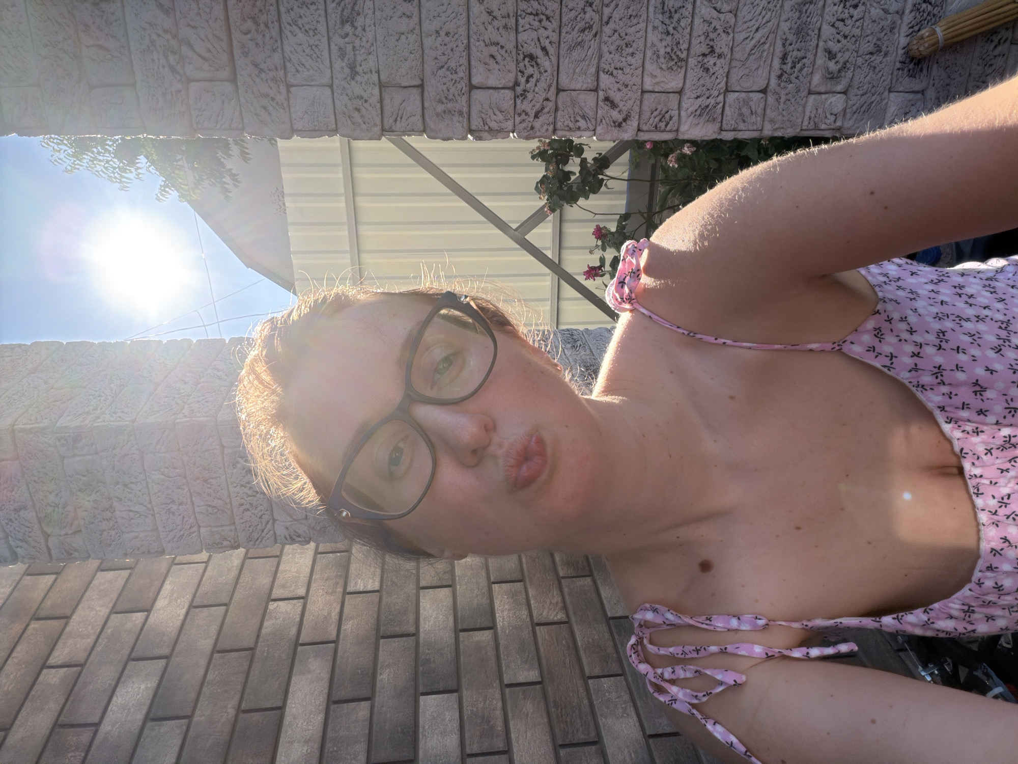A woman with light skin and reddish-blonde hair wears glasses and a pink floral sundress. She puckers her lips for a selfie. The background shows a brick wall, a light-colored fence, and sunlight filtering through trees. The scene appears to be outdoors, possibly in a yard.