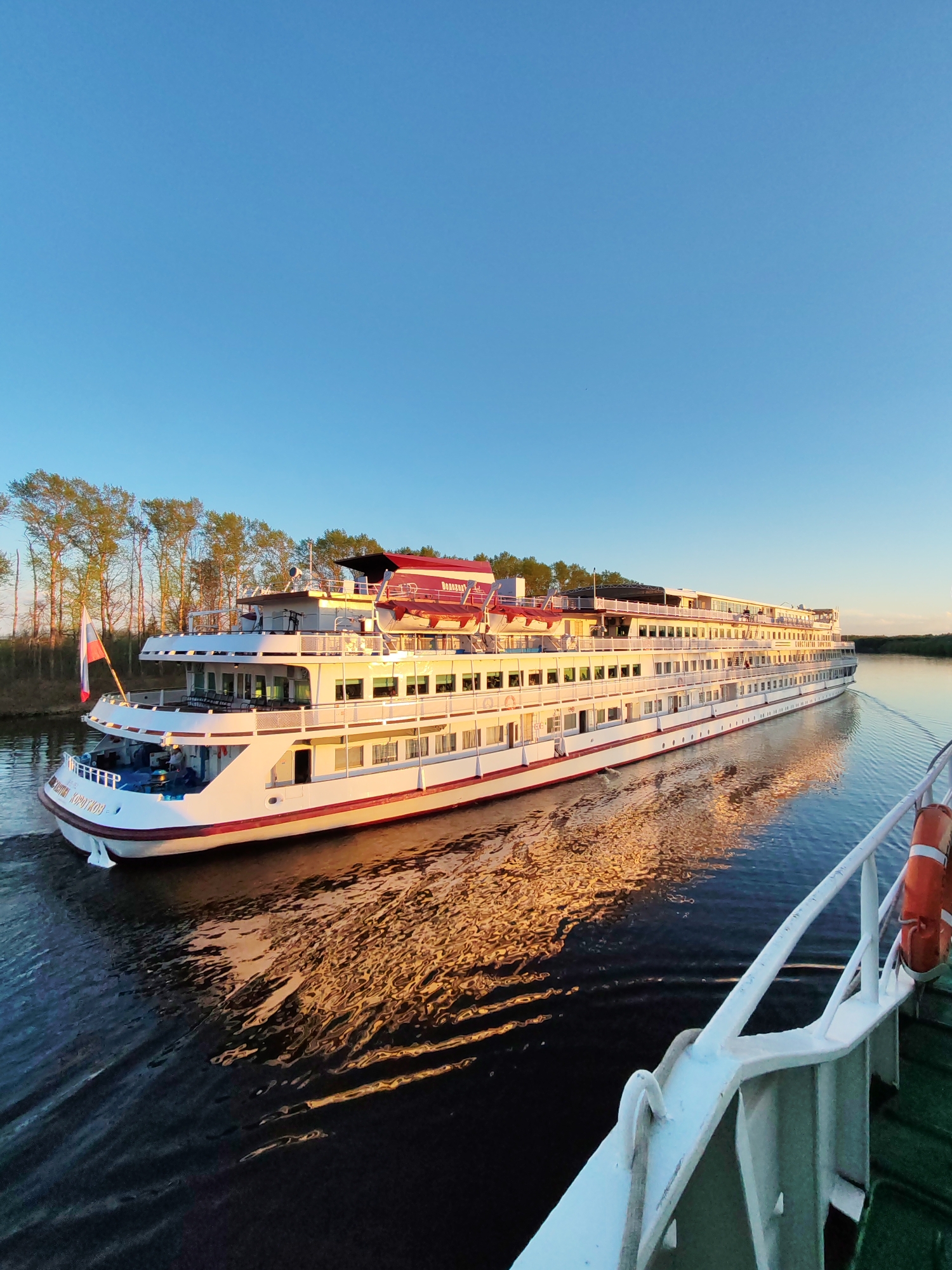 A long, white passenger cruise ship with red accents is moving on a river, its reflection visible in the water. The ship has multiple decks with numerous windows, and the name "Volga Dream" is visible on its side in Russian script. The Russian flag is prominently displayed at the stern.
To the left, a line of trees is visible along the riverbank, illuminated by the setting sun. In the foreground, part of another ship's railing with an orange life preserver is visible. The clear, blue sky takes up the upper half of the frame. The overall scene suggests a river cruise, likely in the vicinity of Myshkin, Russia, given the reference to the Volga Dream, which is known to operate on the Volga River.