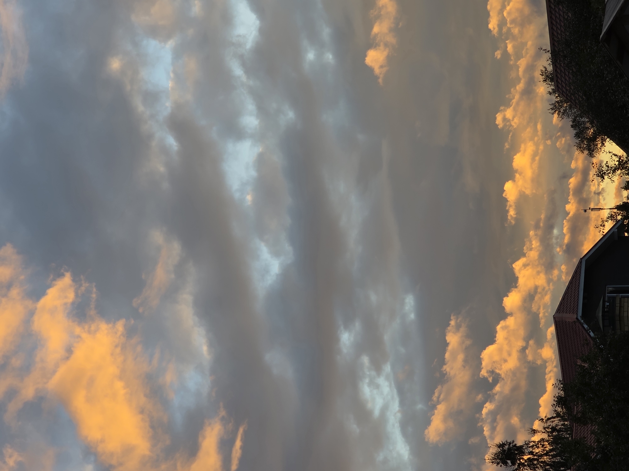 The image shows a dramatic sunset over the city of Rechitsa, Russia. The sky is filled with clouds, some of which are illuminated by the setting sun, casting a warm, orange glow. Other clouds are dark and ominous, suggesting the possibility of rain. In the foreground, the roofs of several houses are visible, silhouetted against the colorful sky. The trees are also dark, their leaves catching the last rays of sunlight. The scene is peaceful and serene, with a sense of the beauty of nature.