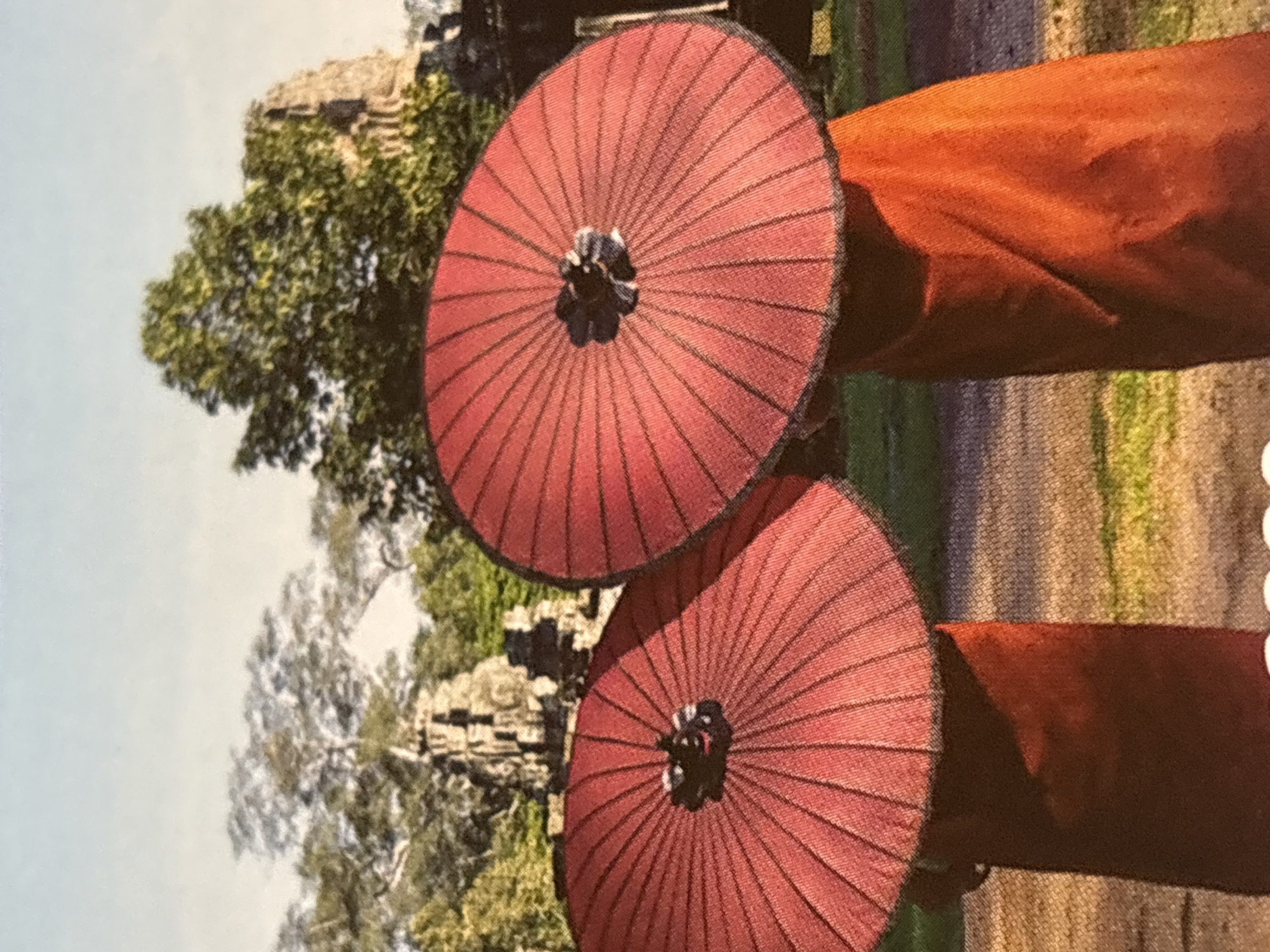 This image depicts two traditional red paper umbrellas, each with a dark central mechanism and thin black ribs radiating outwards. The umbrellas are positioned in the foreground, angled diagonally across the frame. Behind the umbrellas, a blurred background reveals lush green trees and ancient stone ruins, suggesting a historical or temple-like setting. The top portion of the image features a bright, pale blue sky. The lighting appears to be natural, indicating daytime, possibly under clear or partly cloudy conditions. The overall impression is one of serene outdoor beauty, possibly hinting at a cultural or spiritual event, or simply a picturesque scene. There is no visible text in the image.