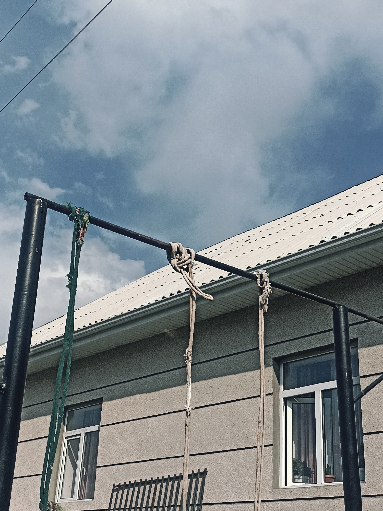 An outdoor, low-angle shot shows a pull-up bar and hanging ropes in front of a building with a white, tiled roof. The pull-up bar is made of black metal and supported by two vertical posts. A green rope is tied to the left side of the bar, and two thicker, beige ropes are tied to the right side. The building has a grey, textured exterior with horizontal lines and two windows. The window on the left has white trim and shows a dark interior. The window on the right also has white trim and is partially open, revealing curtains and houseplants. In the background, the sky is partly cloudy with a blue hue. Electrical wires are visible in the upper left corner of the image. A black metal fence is partially visible in the foreground below the left window. The scene appears to be located in Кош-Дөбө, Kyrgyzstan.