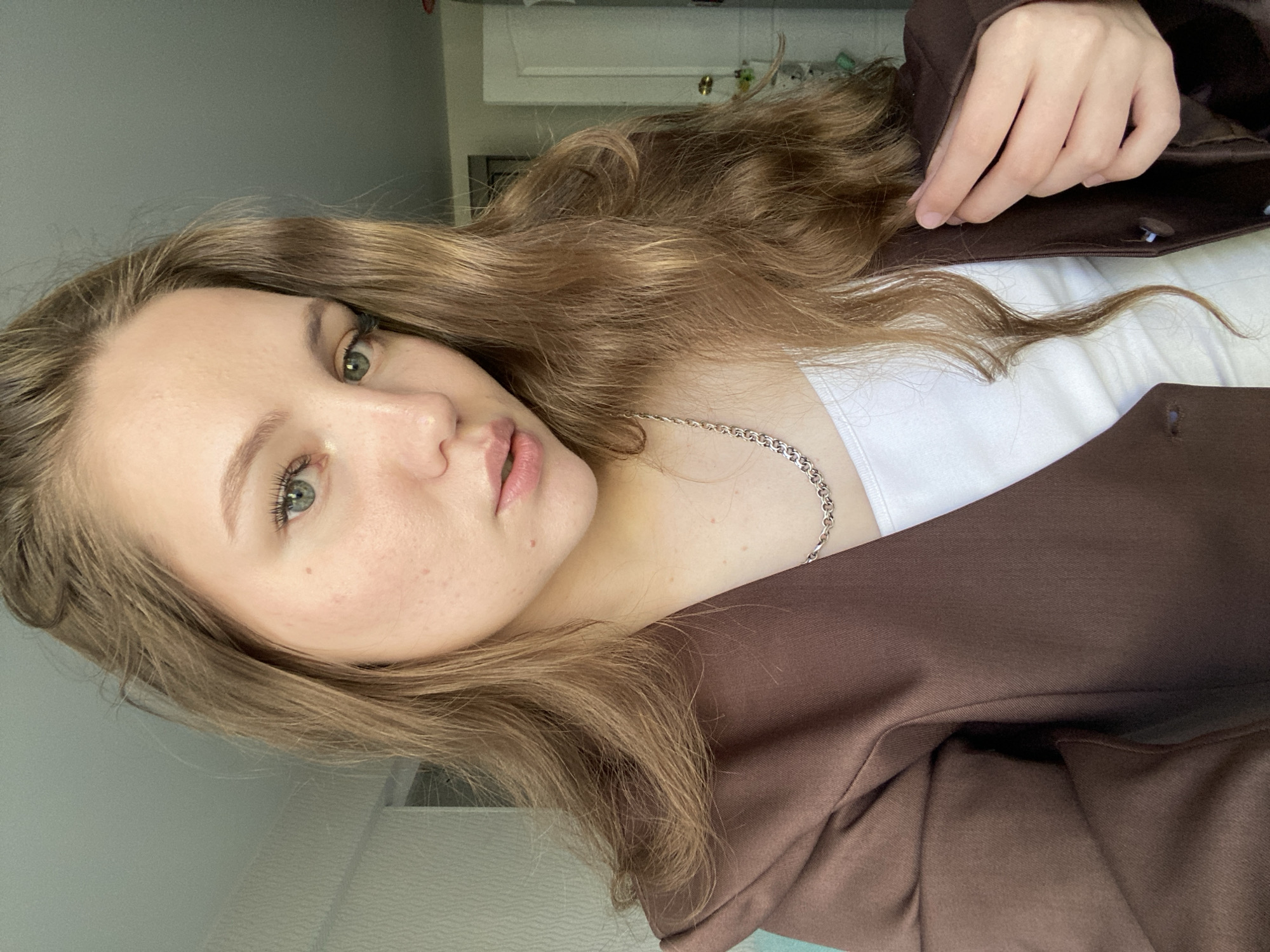 A woman with light brown hair and green eyes is wearing a white top and a brown blazer. She has fair skin and a silver necklace. She is looking directly at the camera. The background is out of focus but appears to be an interior setting. The location in the city of Izhevsk, Russia is unknown.