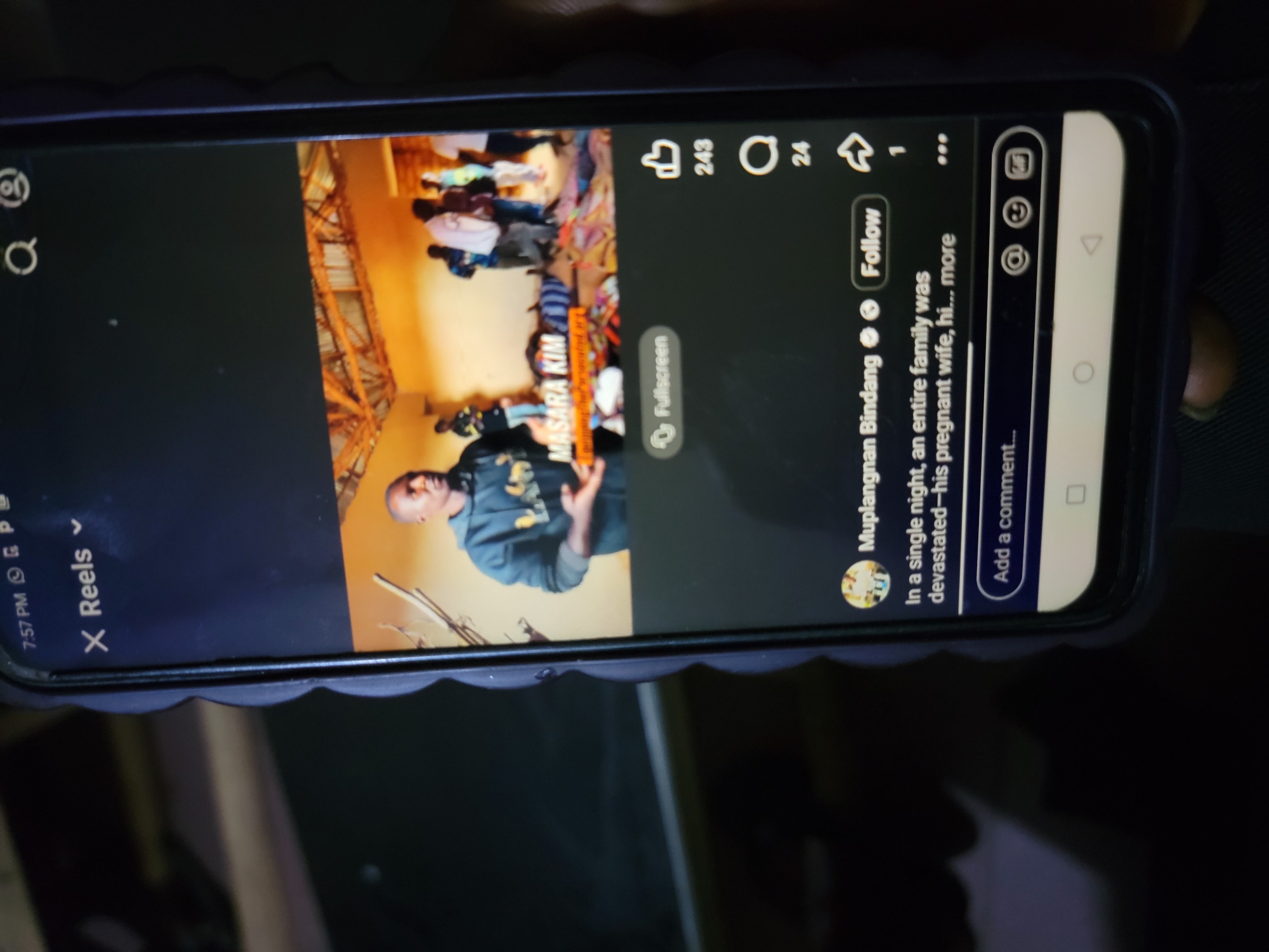 The image shows a smartphone screen displaying a social media reel, possibly on Instagram or TikTok. The reel features a man, identified as Muplangnan Bindang, and a scene of apparent devastation. Text accompanying the reel describes a tragic event involving an entire family, including a pregnant wife. The reel may relate to the location of Masara Kim in Jos, Nigeria. The screen also displays engagement metrics like likes (243), comments (24), and shares (1), with options to follow the user and leave a comment. The time on the phone indicates 7:57 PM. The phone has a protective case and is resting on a dark, cluttered surface.