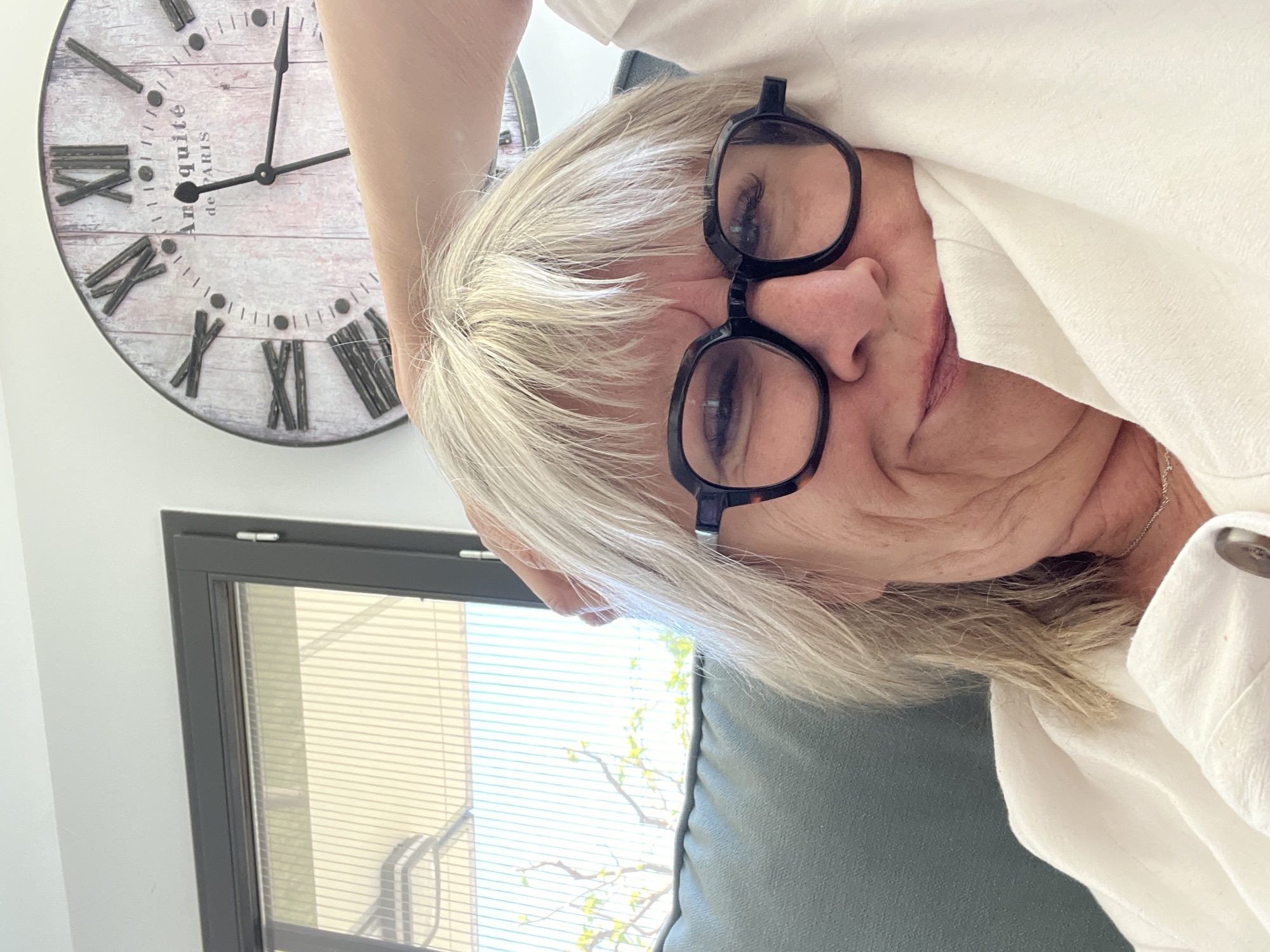 A photograph shows a mature woman with short grey hair and wearing black-rimmed glasses. She is dressed in a white shirt. She appears to be sitting on a grey-blue couch. Behind her is a wall with a large clock displaying Roman numerals and the words "Antiquite de Paris". To the left of the woman, there is a window with blinds. Outside the window, one can see a glimpse of a tree with green leaves. There is no indication that this picture was taken in Chambœuf, France.