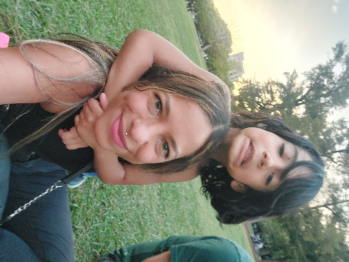 A selfie captures a woman with light brown hair, fair skin, and a lip piercing, sitting in a grassy park. A young girl clings to her back, smiling. The background includes green grass, trees, and a building, suggesting a sunny day. The composition is slightly tilted, emphasizing the informal and candid nature of the photograph.
