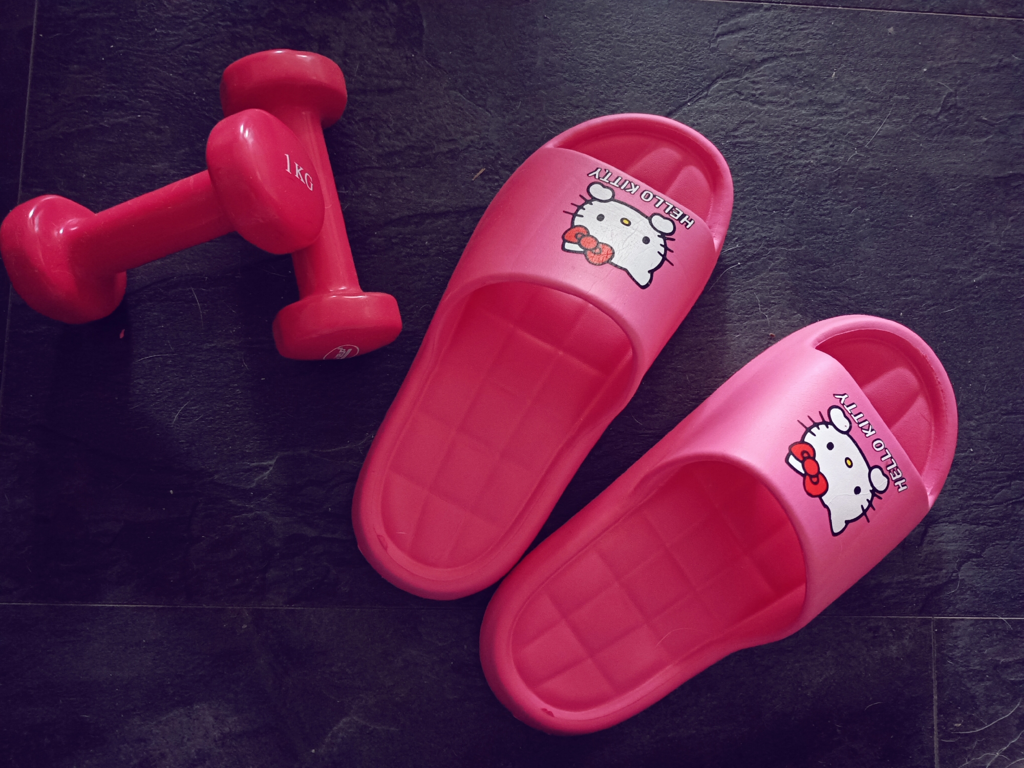 This is a top-down, medium shot of a pair of bright pink Hello Kitty slides and two pink dumbbells on a dark, textured surface that appears to be slate or a similar stone. The dumbbells are positioned to the left of the slides, with one lying horizontally and the other vertically, partially overlapping it. The horizontal dumbbell has "1KG" printed in white on its weight. The slides are arranged diagonally, with the left slide closer to the dumbbells and the right slide positioned further away, towards the bottom right of the frame. Both slides feature the iconic Hello Kitty character with a red bow, and the text "HELLO KITTY" is printed above the character. The lighting suggests it is daytime, with soft shadows. The setting appears to be indoors, given the consistent lighting and the nature of the items displayed, likely a home environment in Sofia, Bulgaria, where exercise equipment and casual footwear are present. There is no discernible activity or event taking place; the items are laid out, suggesting preparation for or completion of an exercise routine, or simply a display of personal items. The overall mood is casual and possibly playful, due to the Hello Kitty motif.