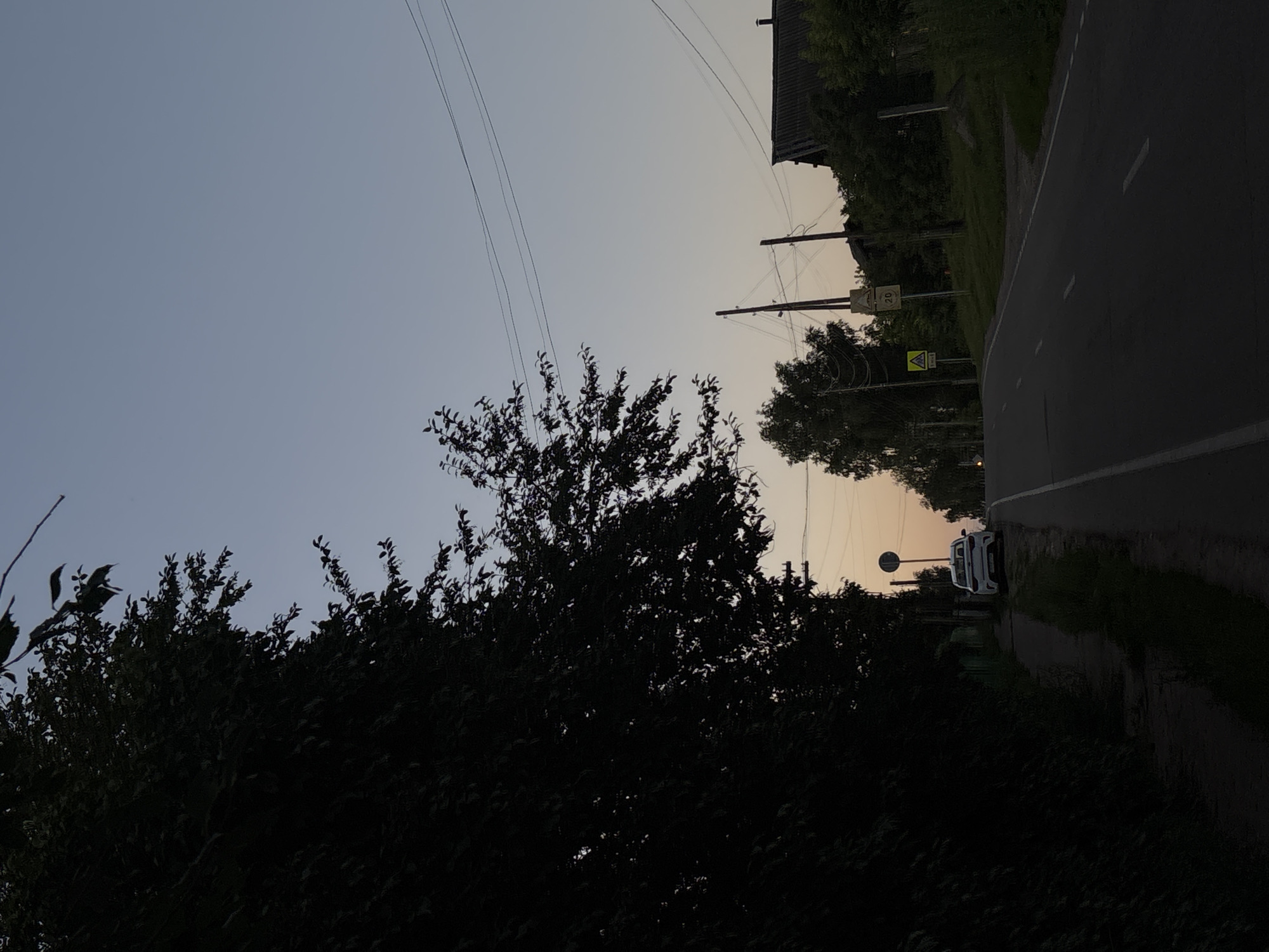 The image depicts a rural road in the city of Fokino, Russia, during twilight. A dark silhouette of dense foliage dominates the left side of the frame, with the tips of branches extending into the pale sky. A white car is parked on the right side of the road, facing away from the camera. The road itself is paved and has visible white lane markings. Power lines are strung across the sky, supported by utility poles. On the right side of the road, a house with a dark roof is visible amongst trees, and a few traffic signs are present, including a yellow triangular sign with a pedestrian symbol and a circular sign with the number "20". The sky transitions from a pale blue at the top to a warmer, hazy orange near the horizon, suggesting the end of a day. The overall atmosphere is tranquil and somewhat muted due to the low light conditions.