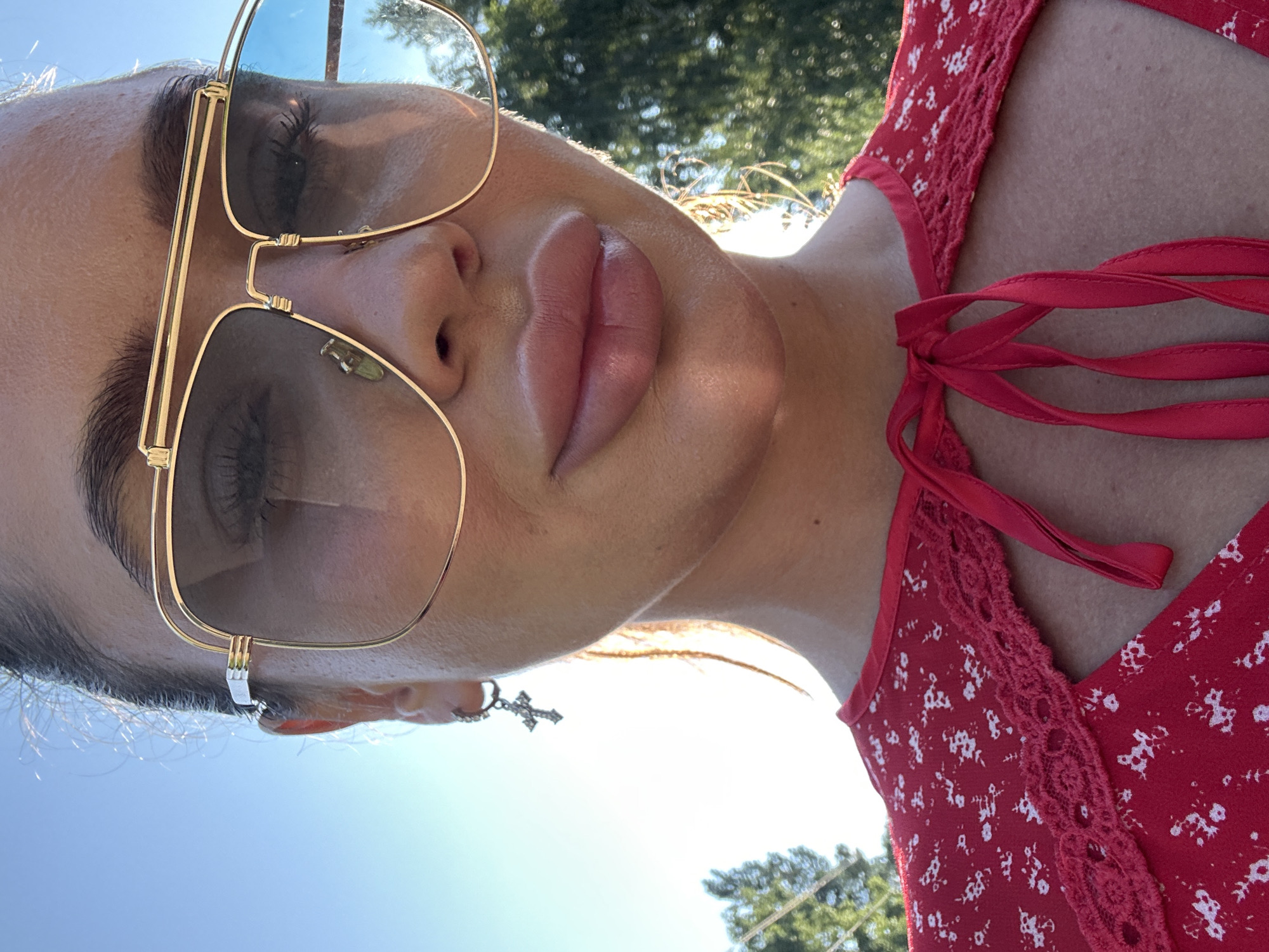 A close-up, eye-level, medium outdoor shot shows the upper body of a woman. She is wearing large, gold-rimmed aviator sunglasses with blue-tinted lenses, a red dress with a white floral pattern and a lace trim, and a cross earring on her left ear. The woman's face is illuminated by natural sunlight. Behind her, the sky is clear and blue, and blurred green trees are visible. The scene is set in Duvankul', Russia.