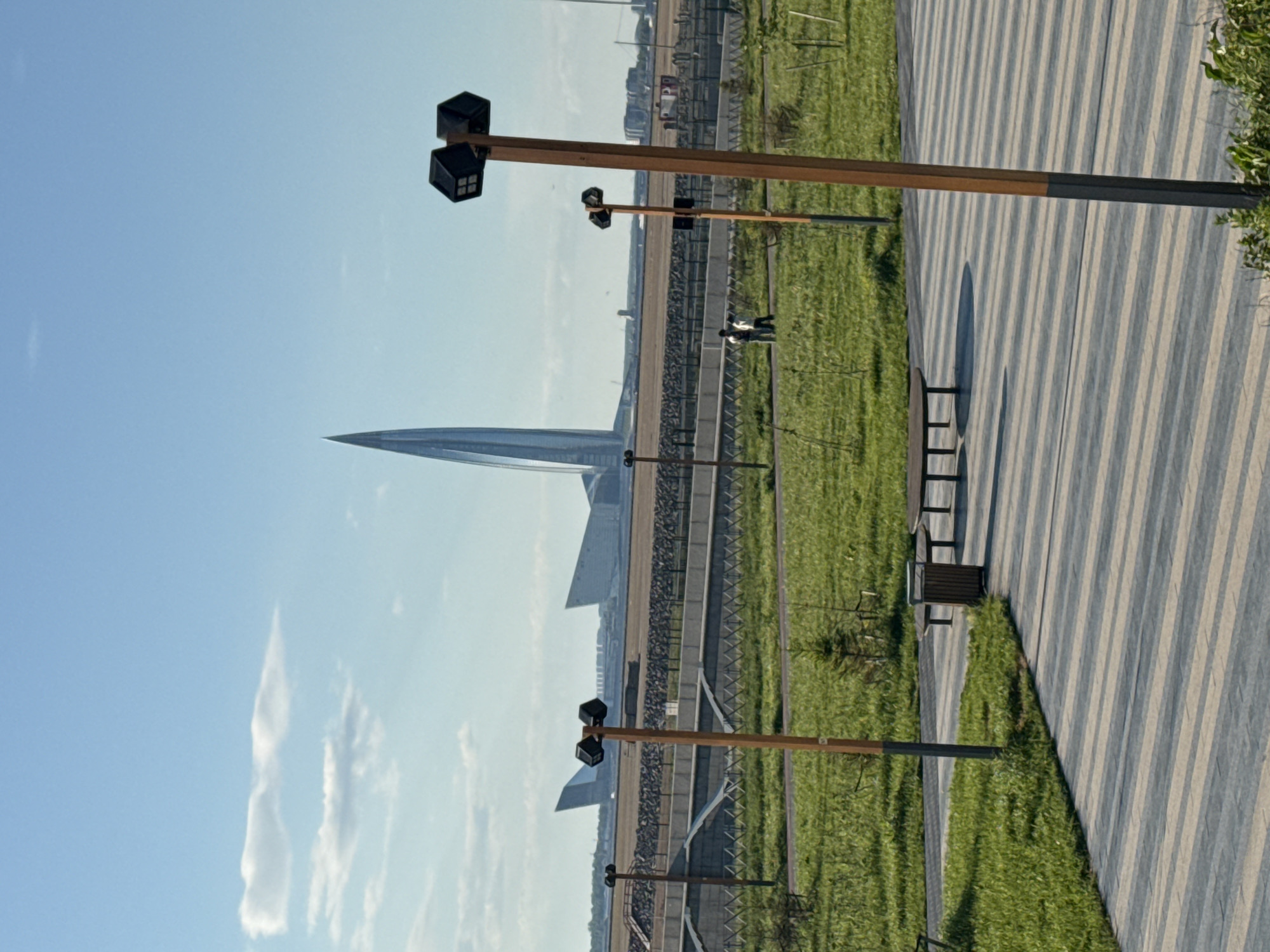 The image depicts a scene in Saint Petersburg, Russia, featuring the Lakhta Center skyscraper. The skyscraper, characterized by its pointed, spire-like design, is seen in the background against a clear blue sky with some scattered clouds. The foreground consists of a park-like setting with green grass and a striped walkway. Several tall light posts with cube-shaped lamps are visible along the walkway. Benches are placed along the walkway for seating. A couple of people are walking in the distance. A fence separates the grass and gravel pathways.