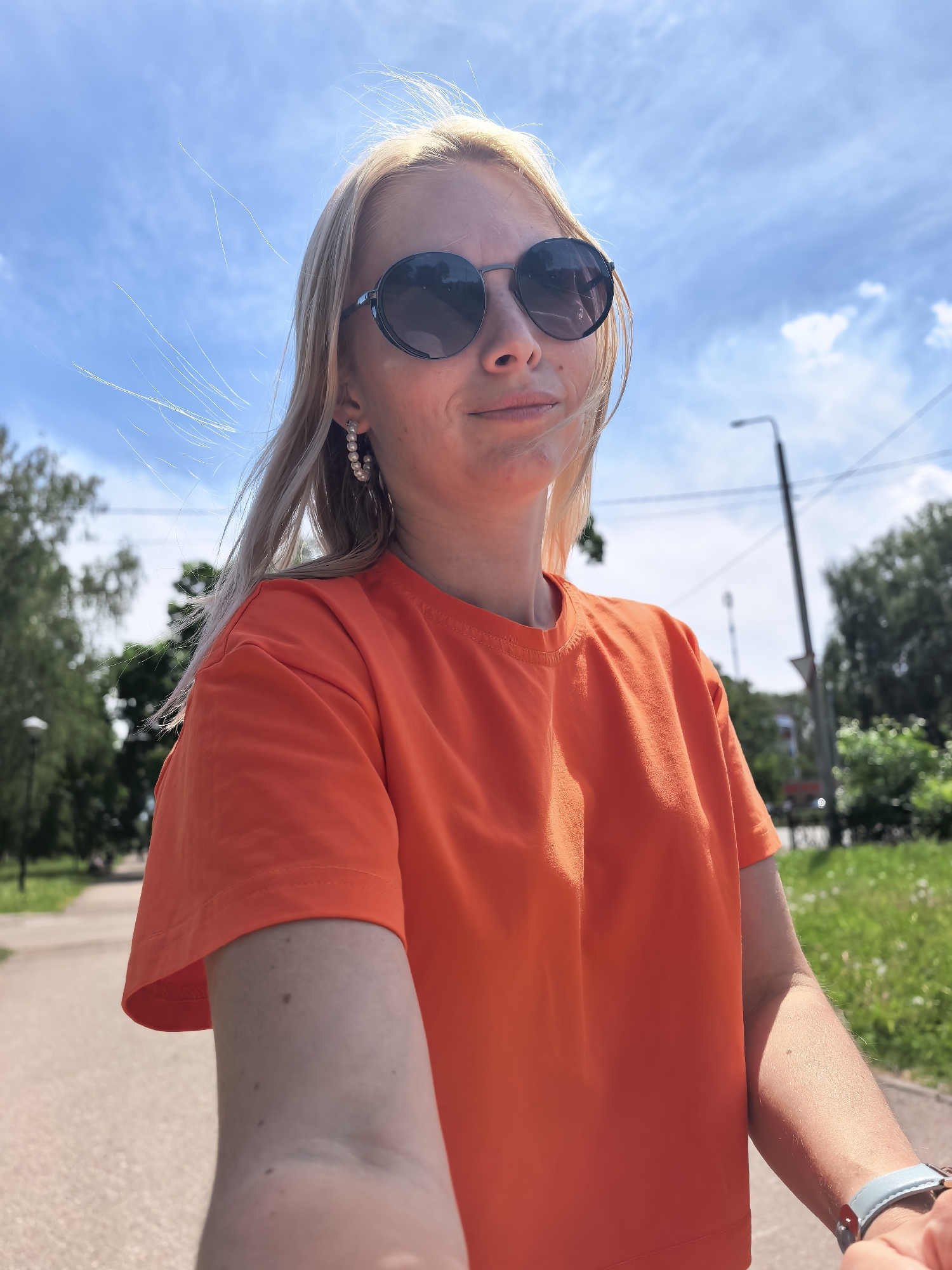 A woman with blonde hair, wearing round sunglasses and pearl earrings, is taking a selfie outdoors on a sunny day in Sterlitamak, Russia. She is wearing an orange T-shirt. In the background, there is a walkway, trees, and a blue sky with scattered clouds. A street lamp is visible.