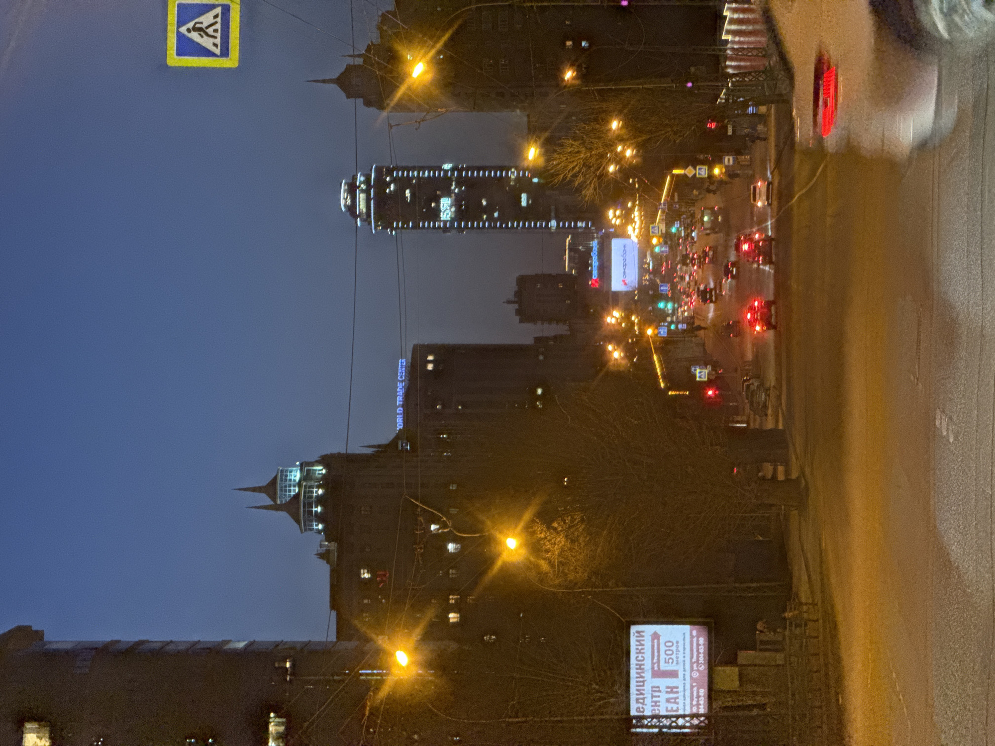 The image shows a nighttime view of Yekaterinburg, Russia, from a street-level perspective. The street is wet and reflecting light. Streetlights and building lights illuminate the scene, showing dense traffic with visible headlights and taillights. Prominent buildings, including a modern skyscraper with decorative lighting, are visible in the background against a dark blue sky. A pedestrian crossing sign is present in the upper left corner. At the bottom of the image, a billboard with Cyrillic text is visible near a sidewalk.